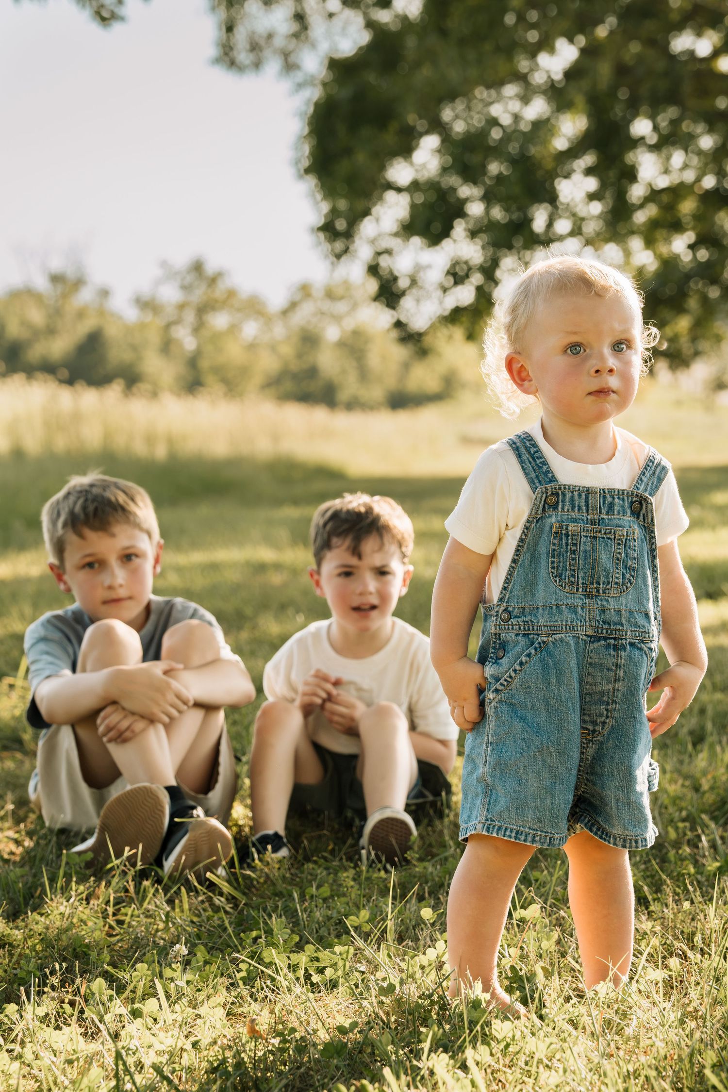 Three young children playing outdoors in a sunny green field, with the youngest girl in denim overalls standing in front.