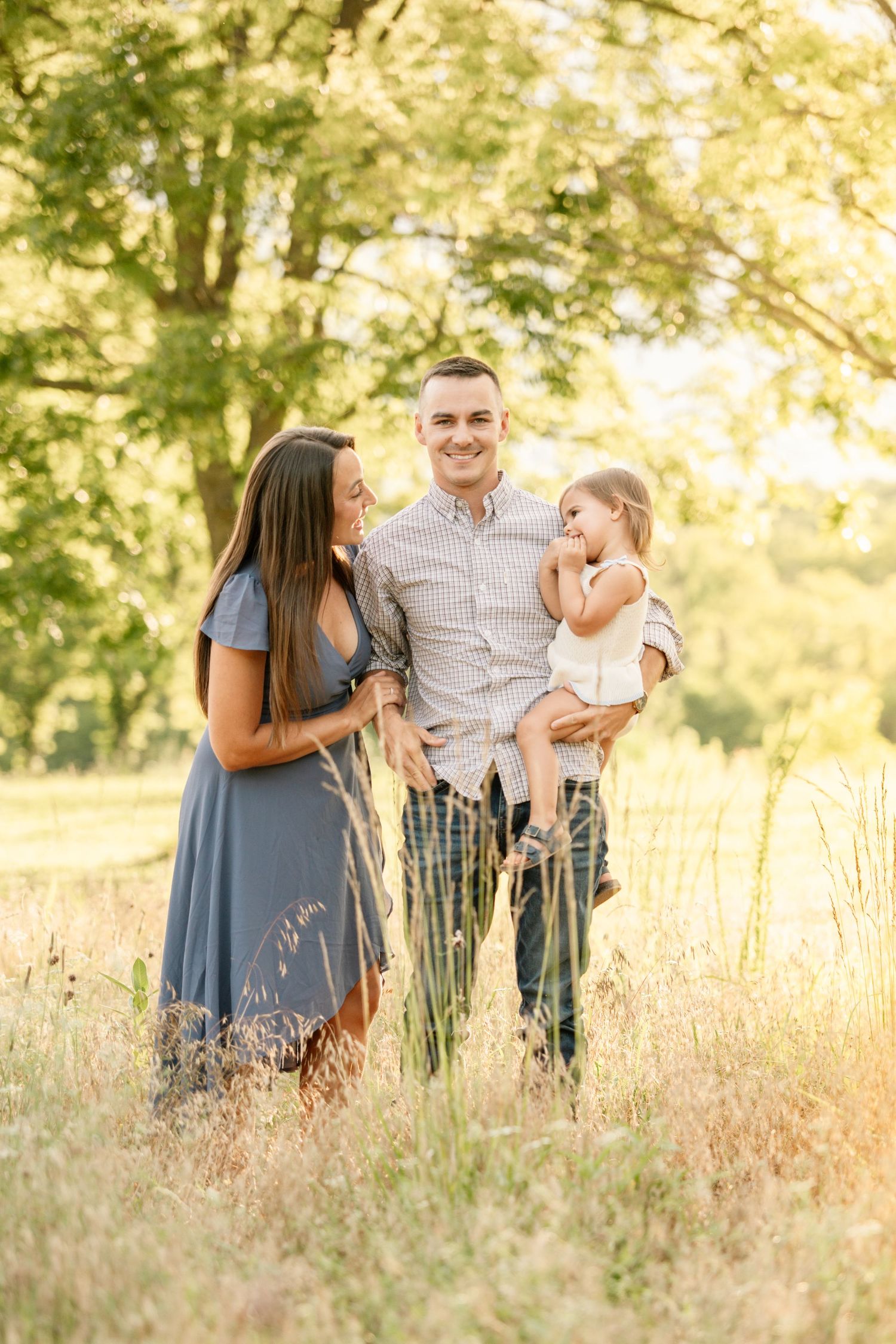 Happy young family with toddler daughter posing in golden sunlit meadow with tall grass.