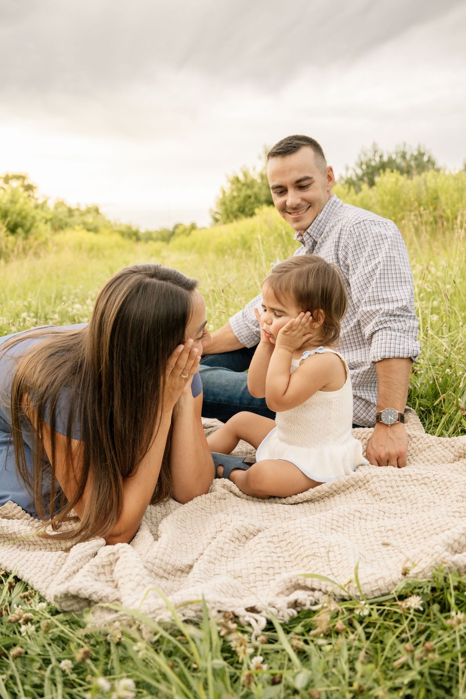 Family sitting on a blanket in a meadow playing peek-a-boo with their toddler daughter outdoors.