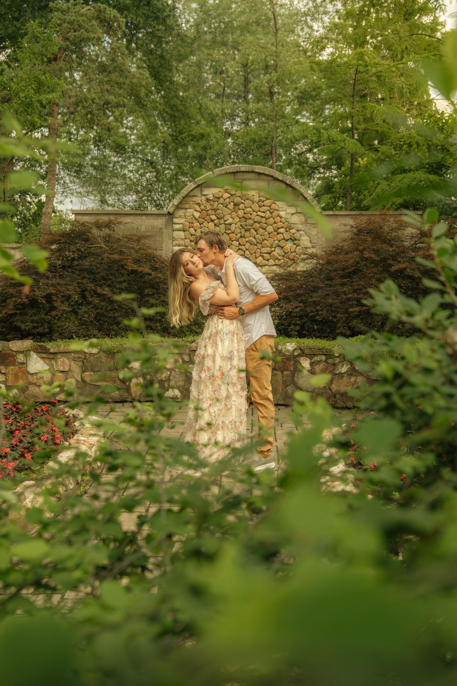 Couple embracing and kissing in a lush garden with a stone arch wall backdrop.
