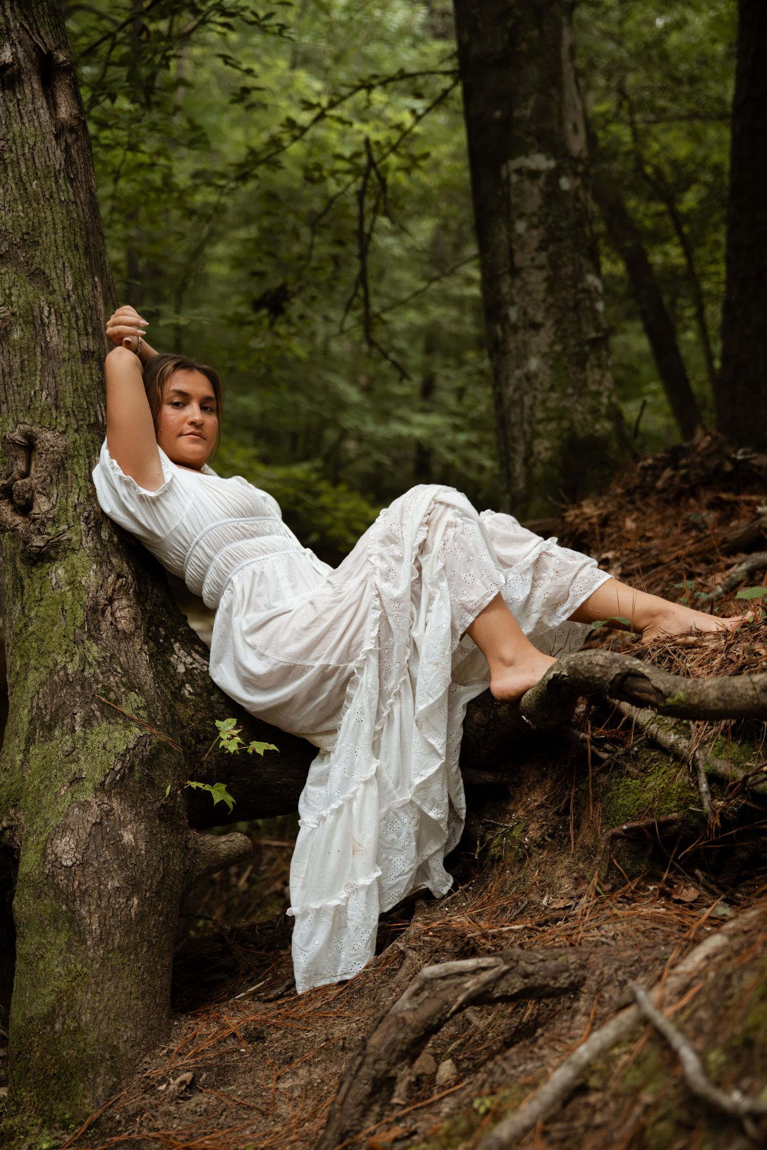Woman in white dress reclining gracefully against tree roots in a lush green forest setting.