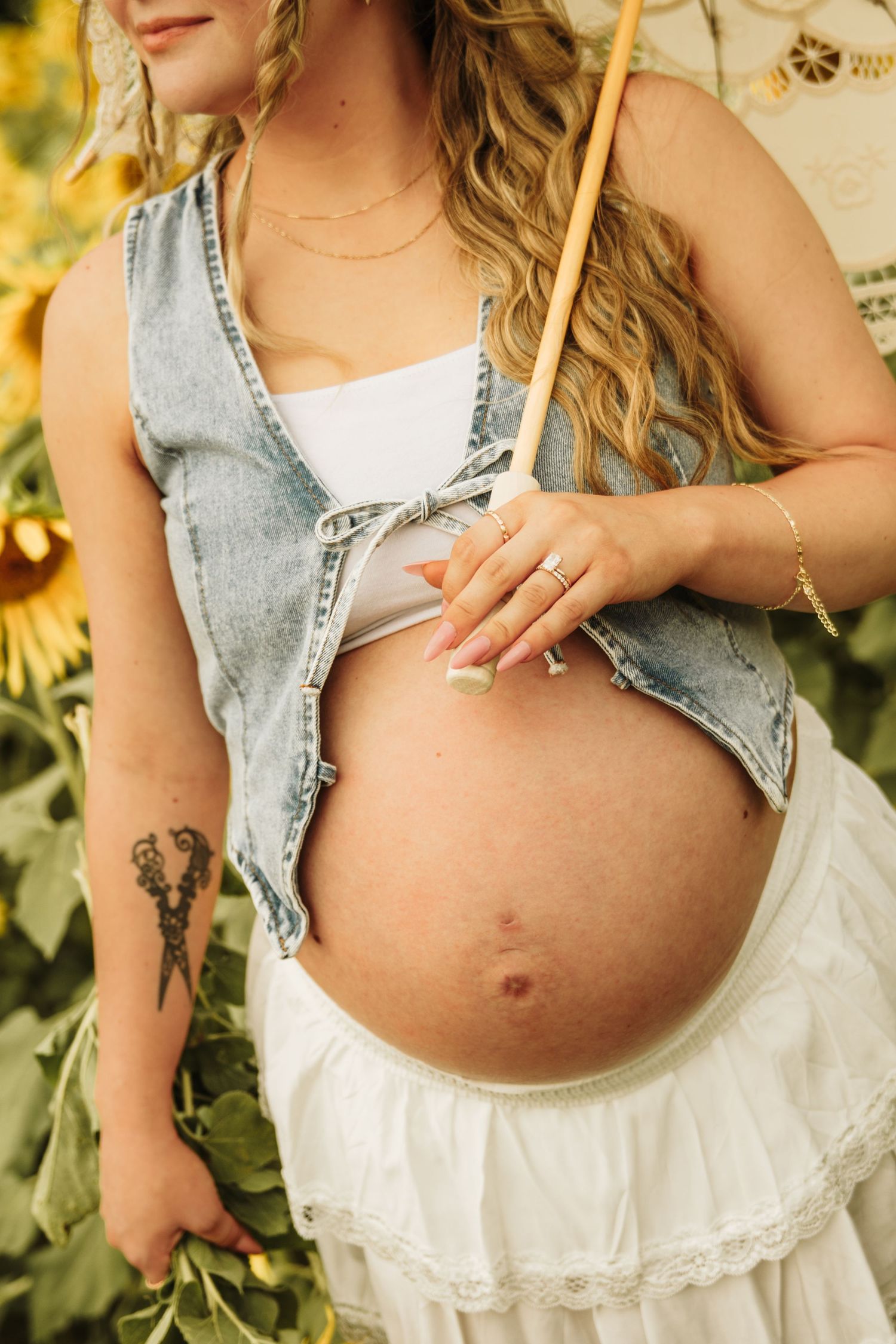 Pregnant woman in denim vest and white skirt holds her bare baby bump in a sunflower field.