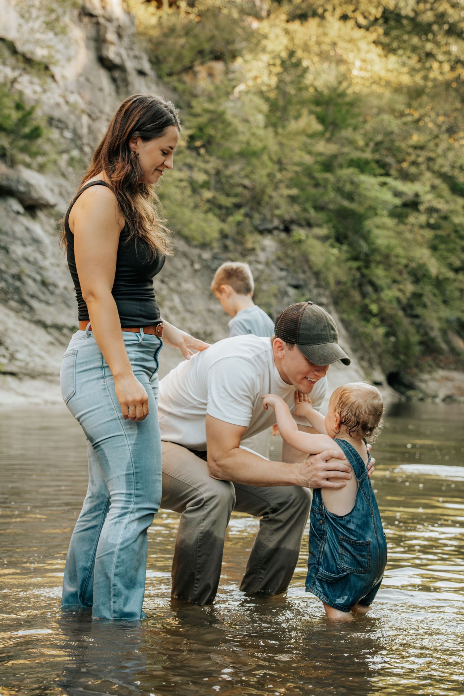 Family wading in a shallow river, father kissing young child while mother smiles, older child visible in background near rocky cliff.