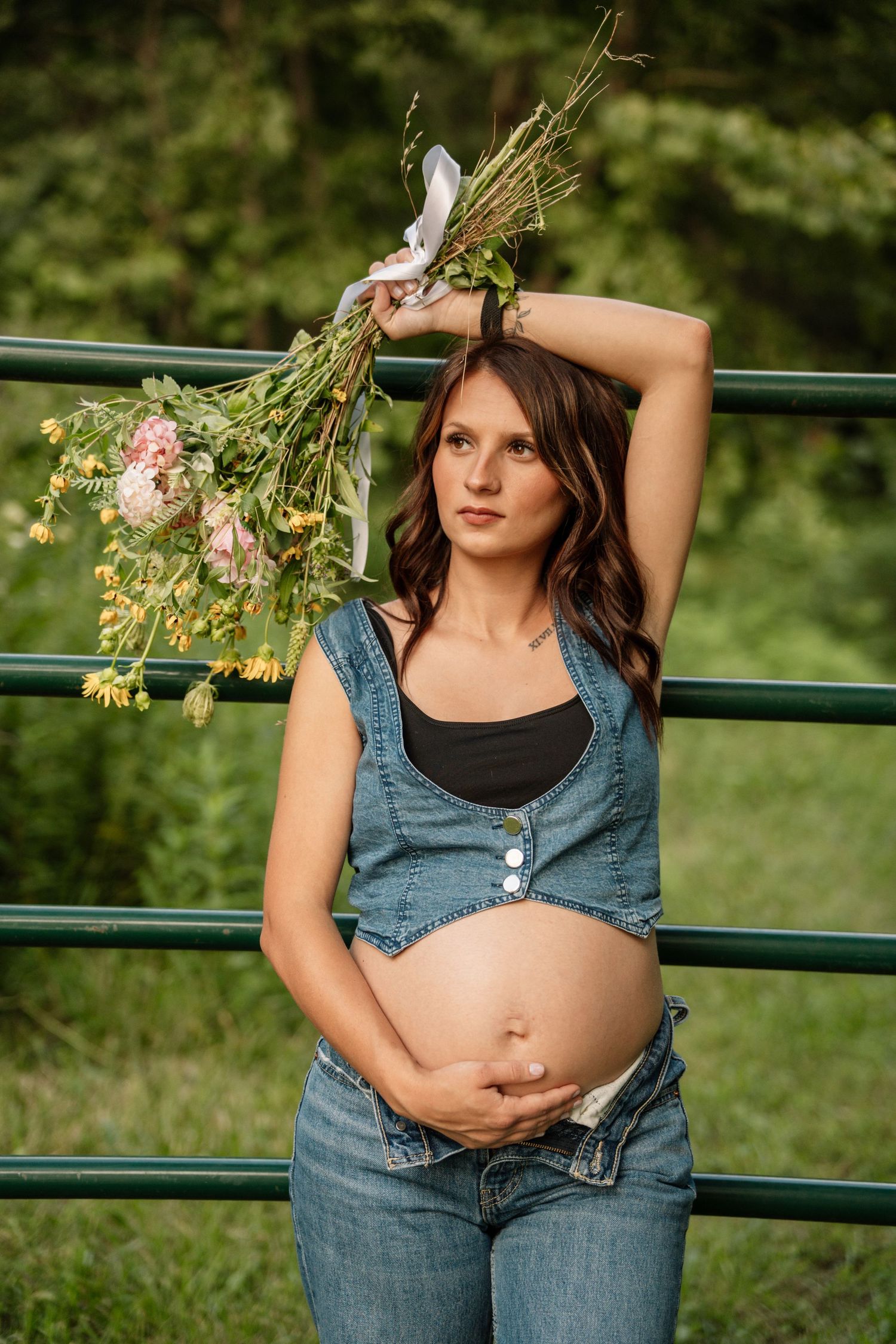Pregnant woman in denim overalls holding wildflowers overhead while leaning on green metal fence outdoors.
