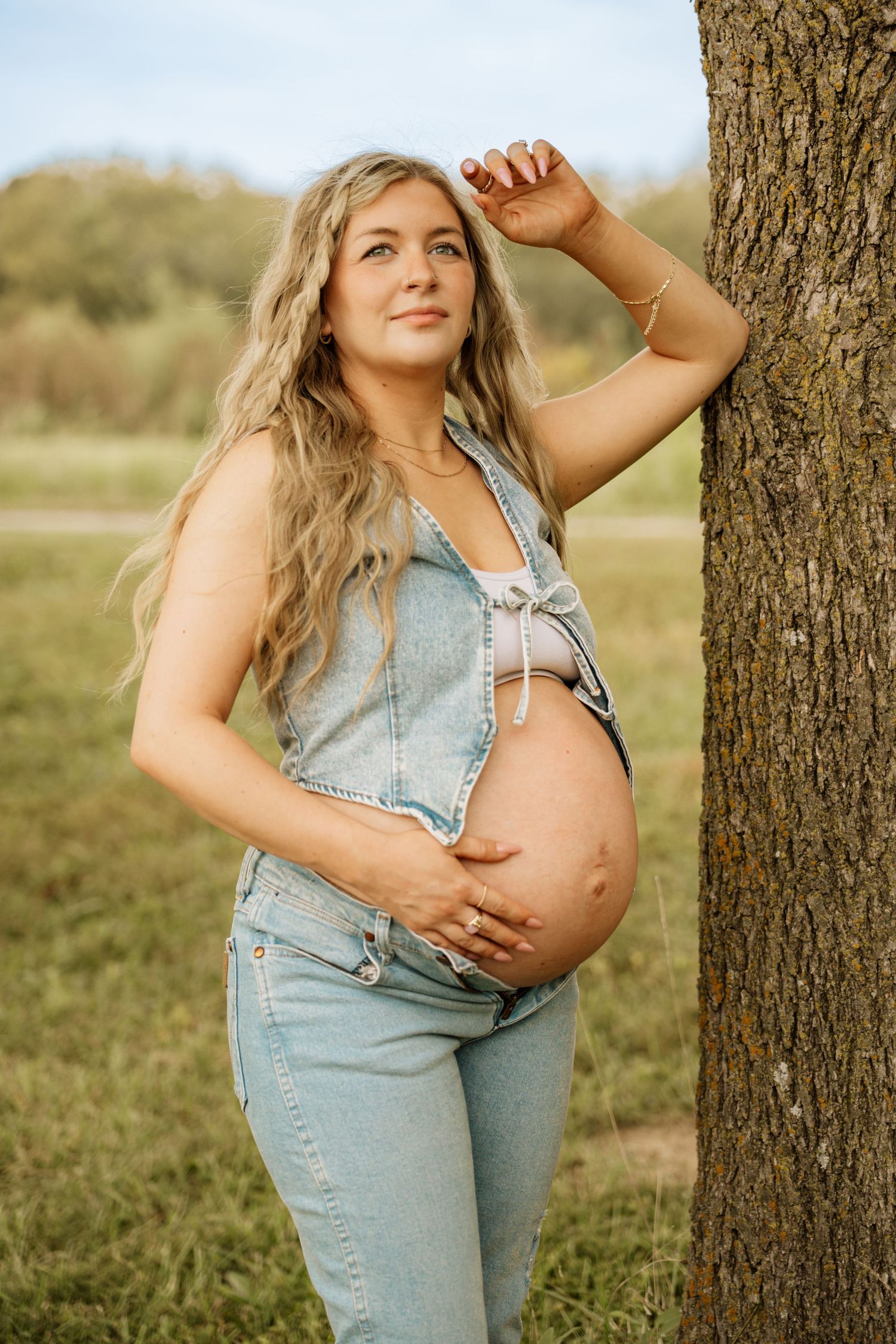 Pregnant blonde woman in denim overalls leans against a tree trunk in a sunny outdoor field.