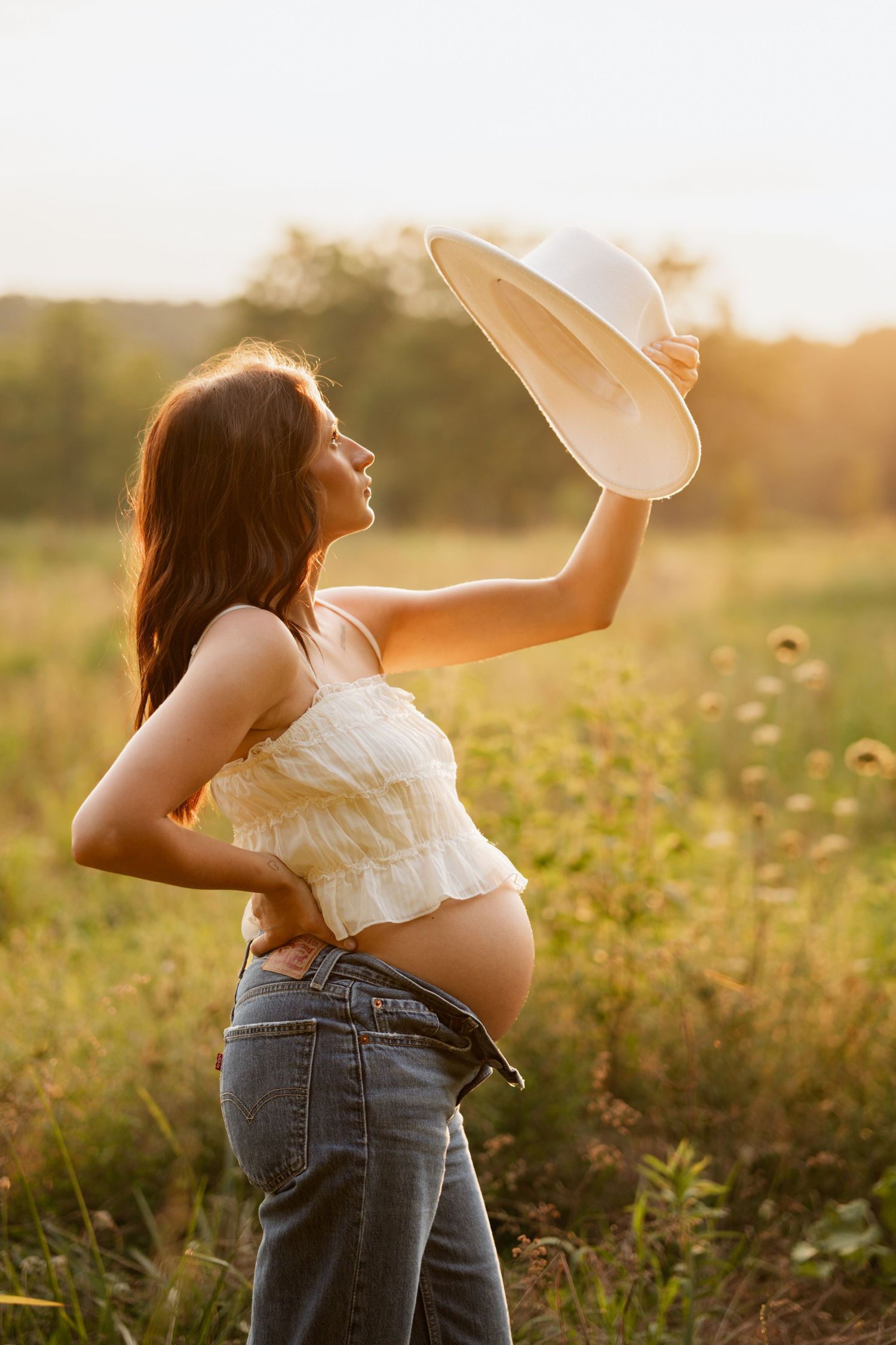 Smiling pregnant woman in white crop top and jeans lifts white hat in sunny golden field.