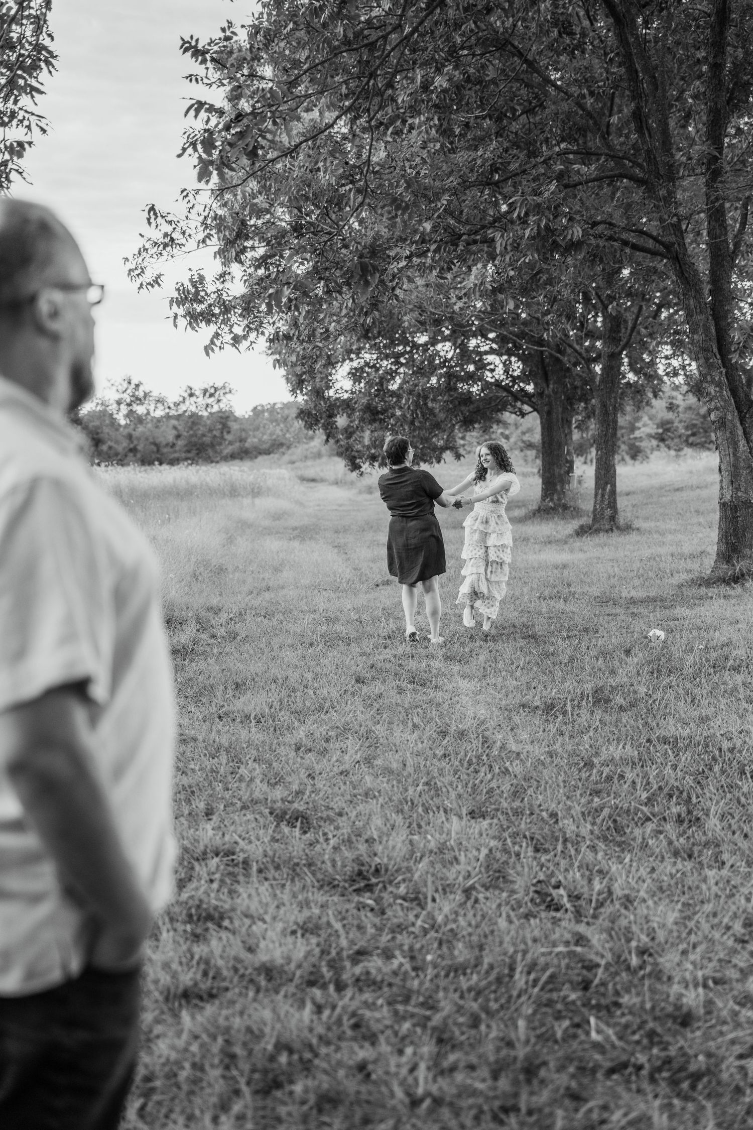 Black and white photo of people gathering under trees in a grassy field, with a man standing in the foreground.