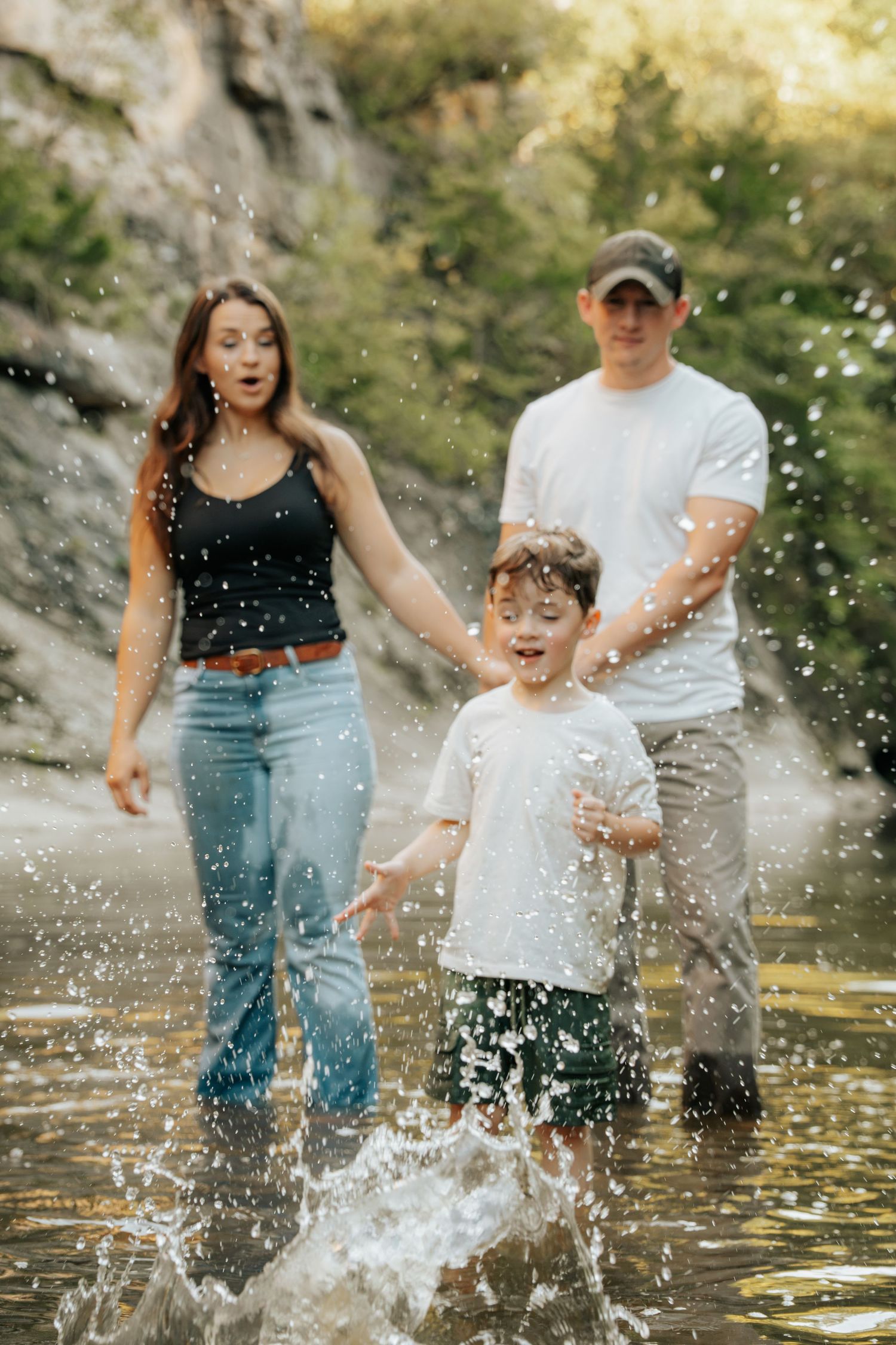 Family of three splashing in a rocky creek, parents holding child's hands outdoors.