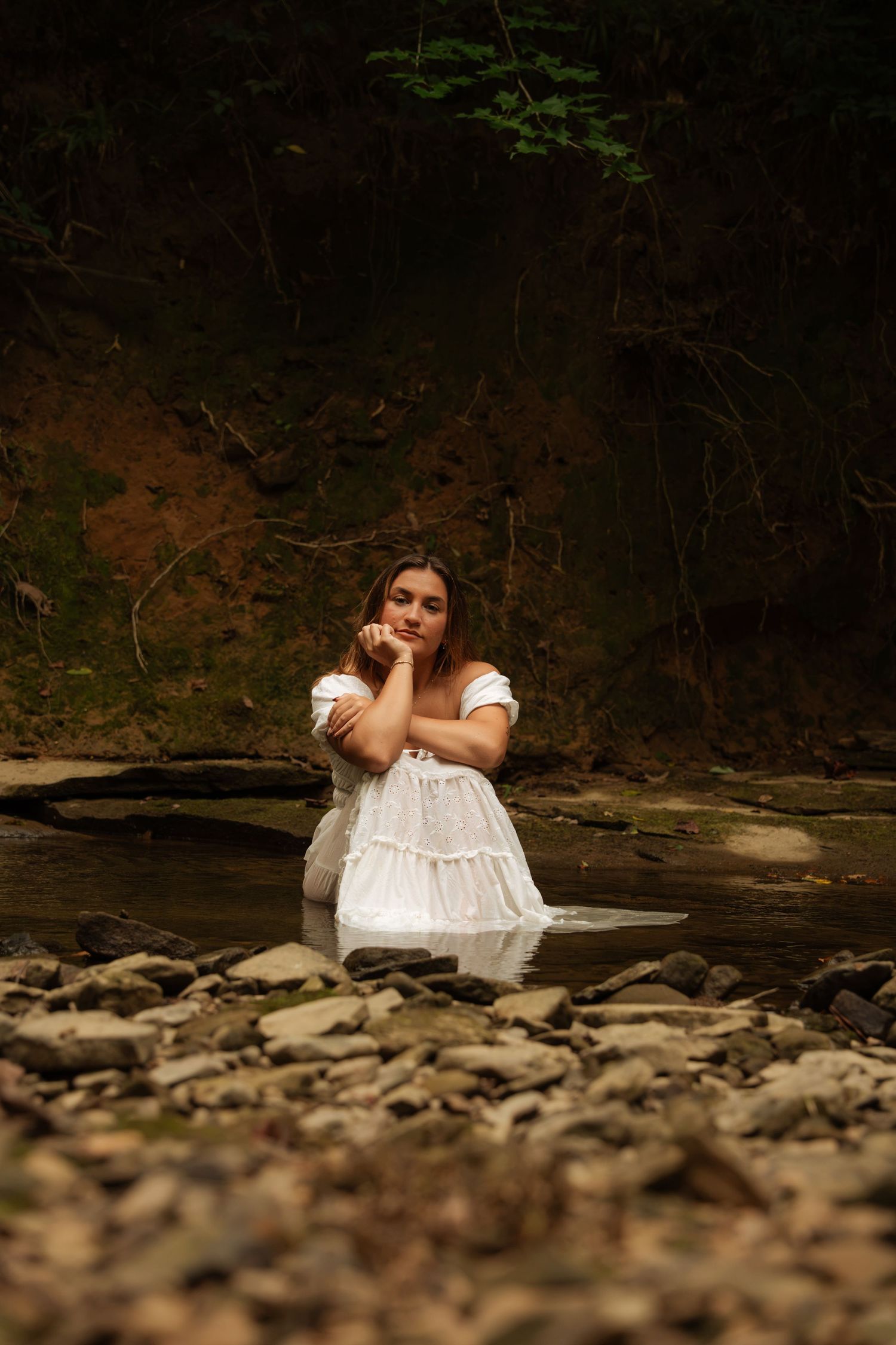 Woman in white dress sitting in rocky stream surrounded by dark forest.