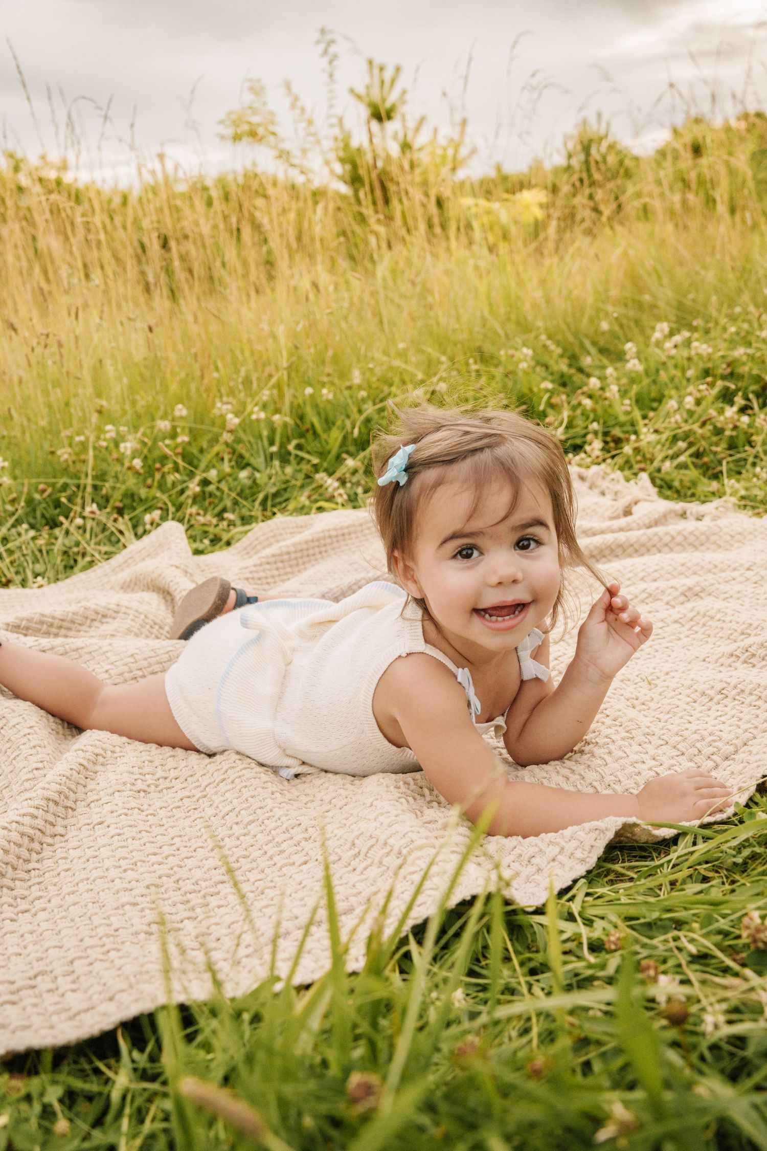 A happy baby girl in a white dress lies on a knit blanket in a sunny green meadow, smiling at the camera.