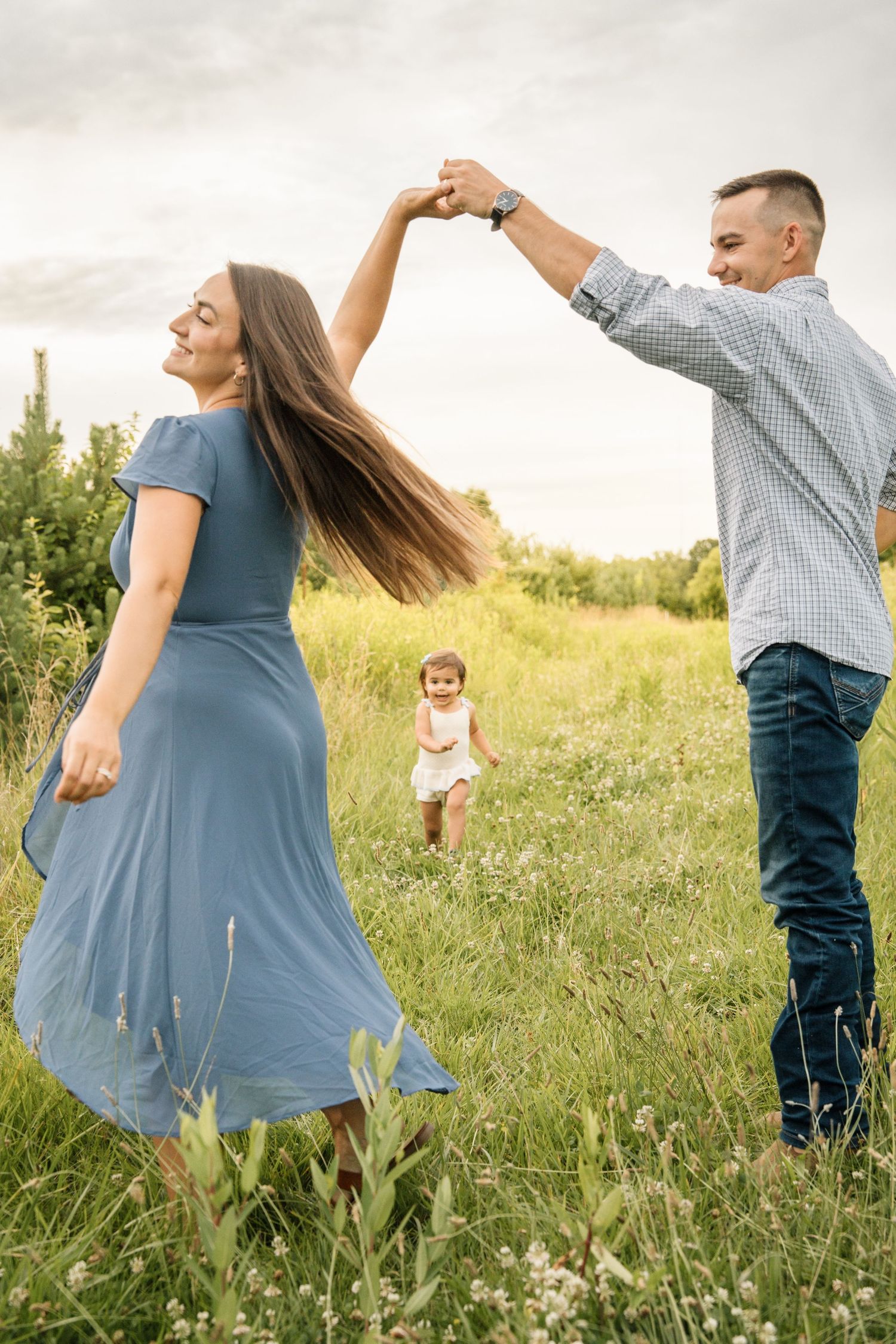 Man spinning woman in blue dress in green meadow while toddler runs toward them in background.