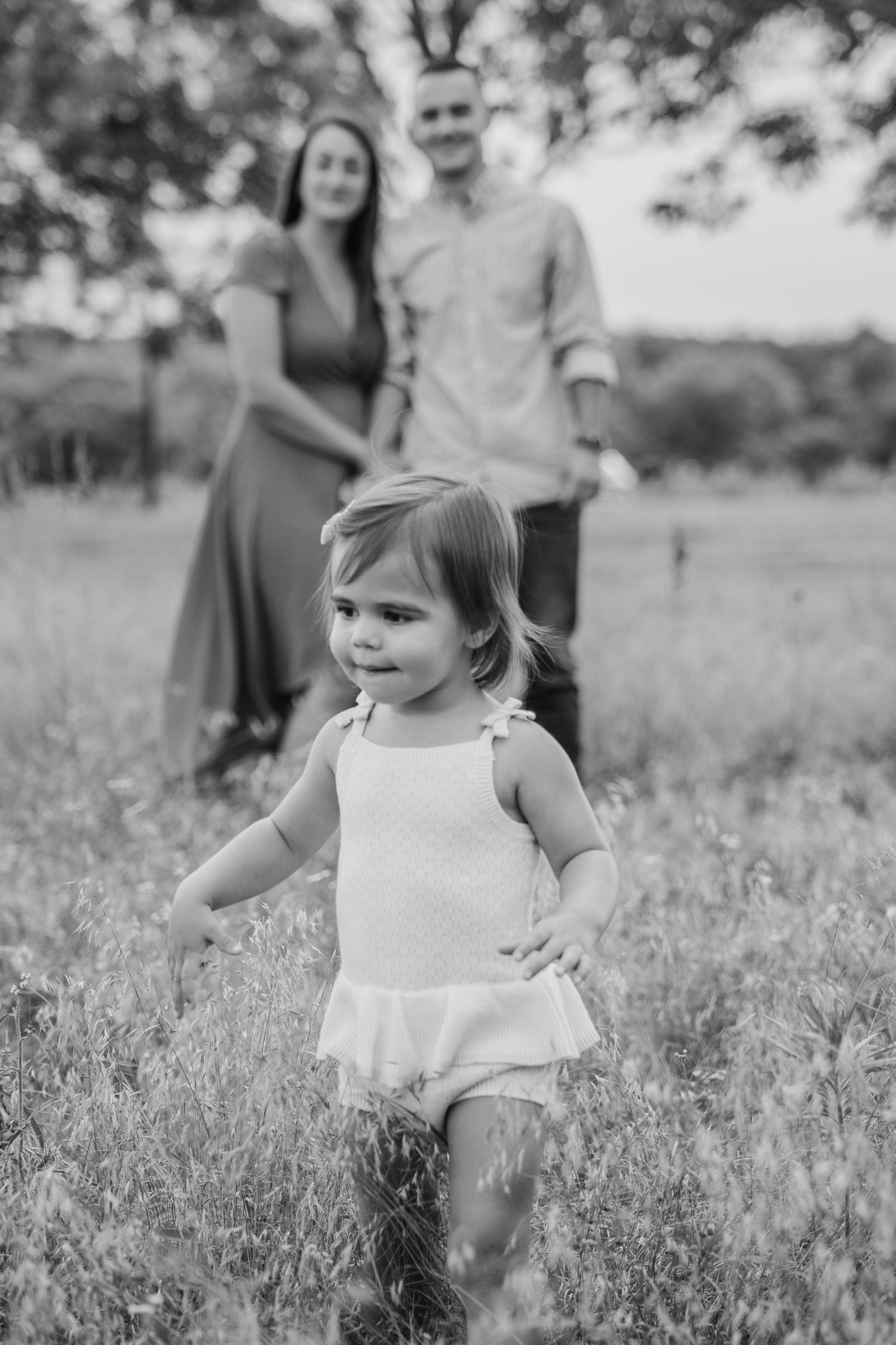 Black and white family photo of toddler girl running in field with parents standing behind her.
