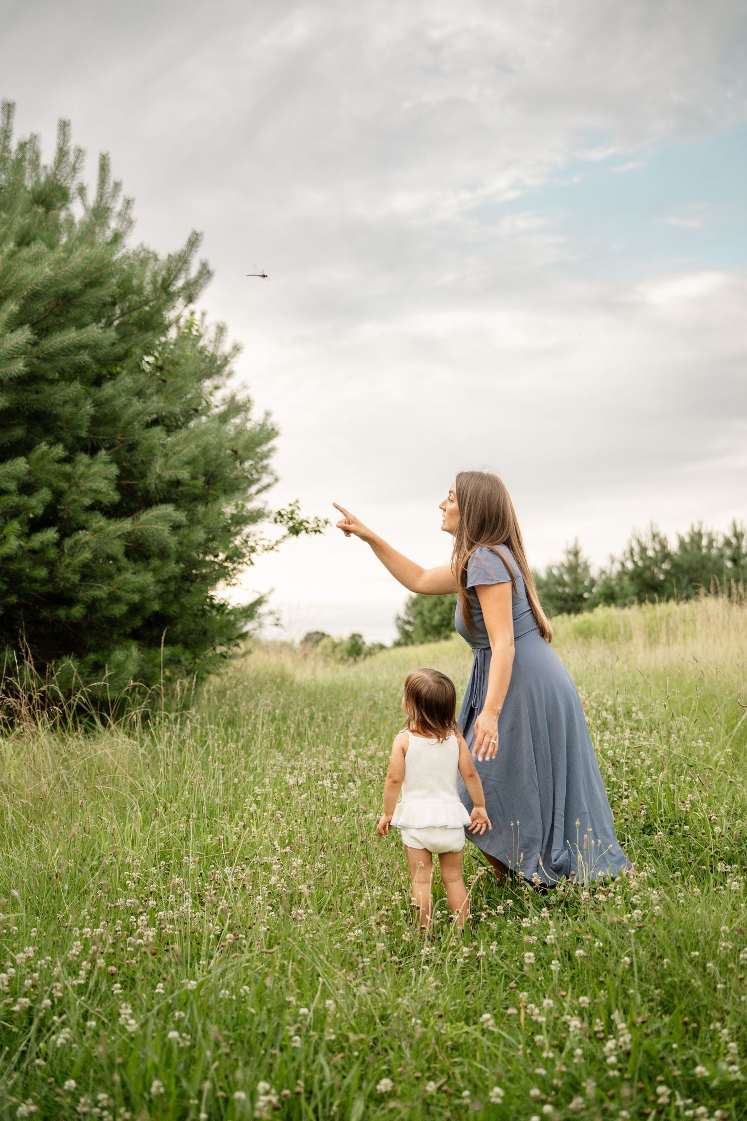 Mother and toddler daughter standing in green meadow, woman pointing at sky while holding wildflowers on cloudy day.