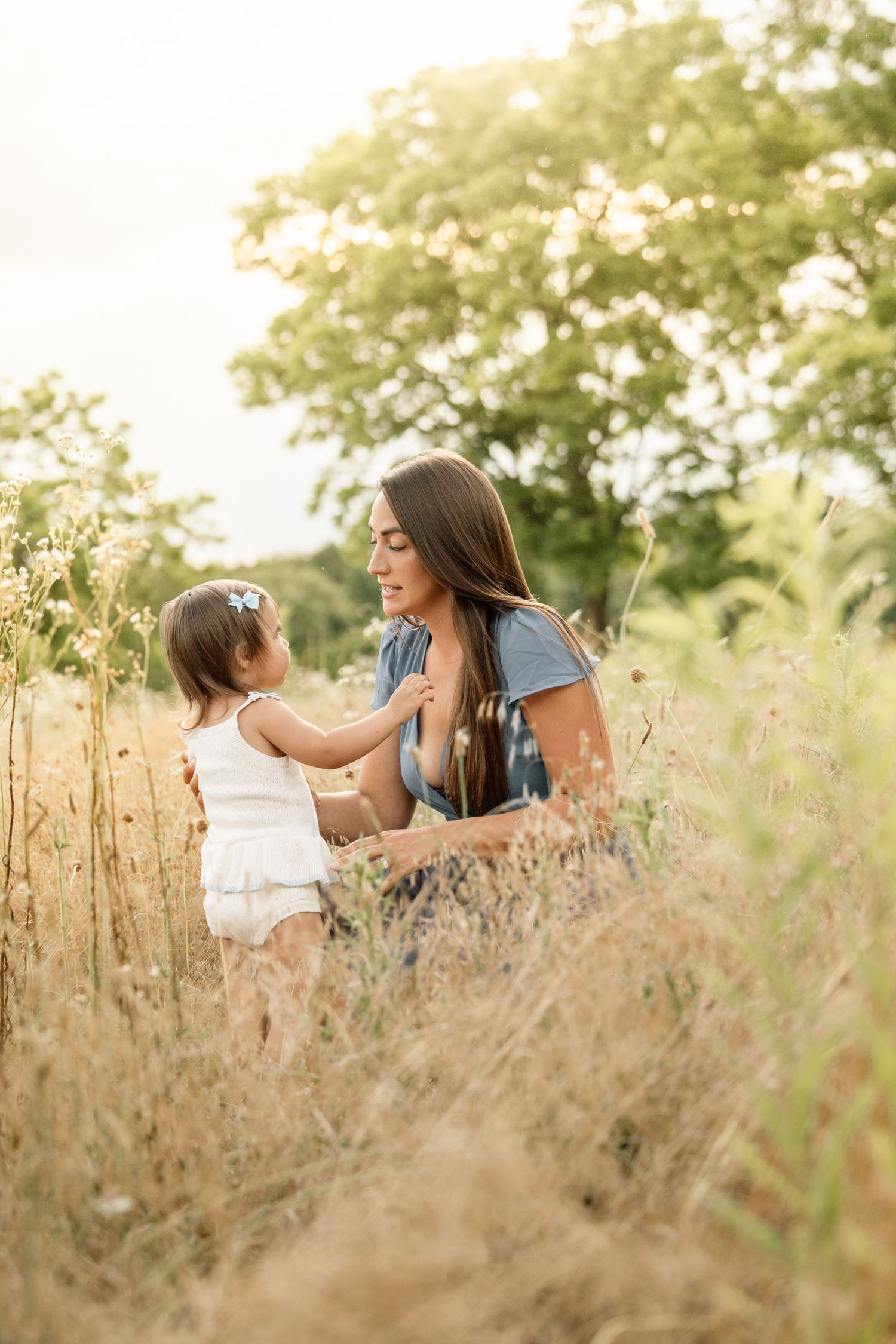 Mother kneeling in golden field interacting tenderly with her young toddler daughter outdoors.