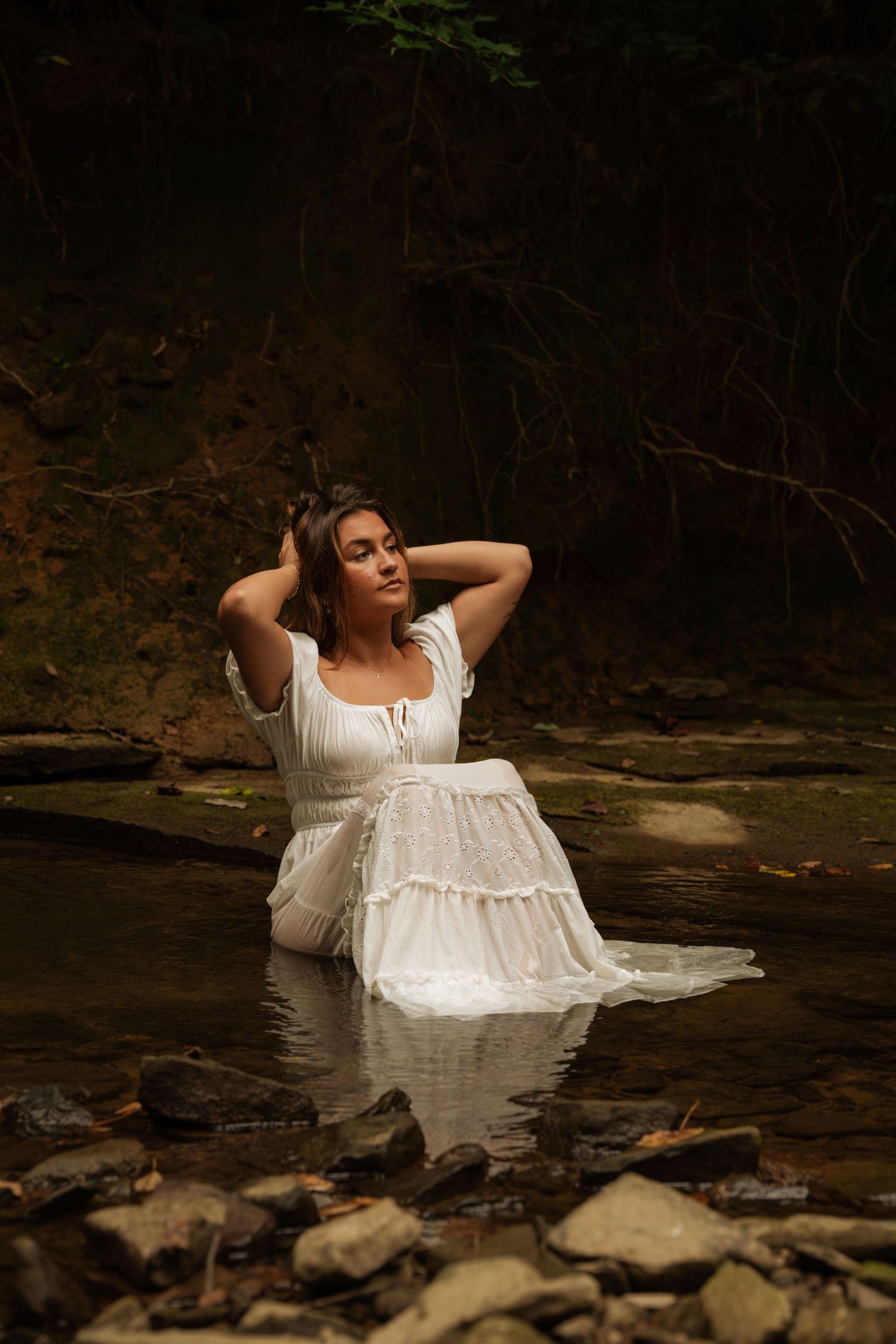 Woman in white dress sitting in a shallow forest stream, hands in hair, surrounded by rocks and dark woodland.