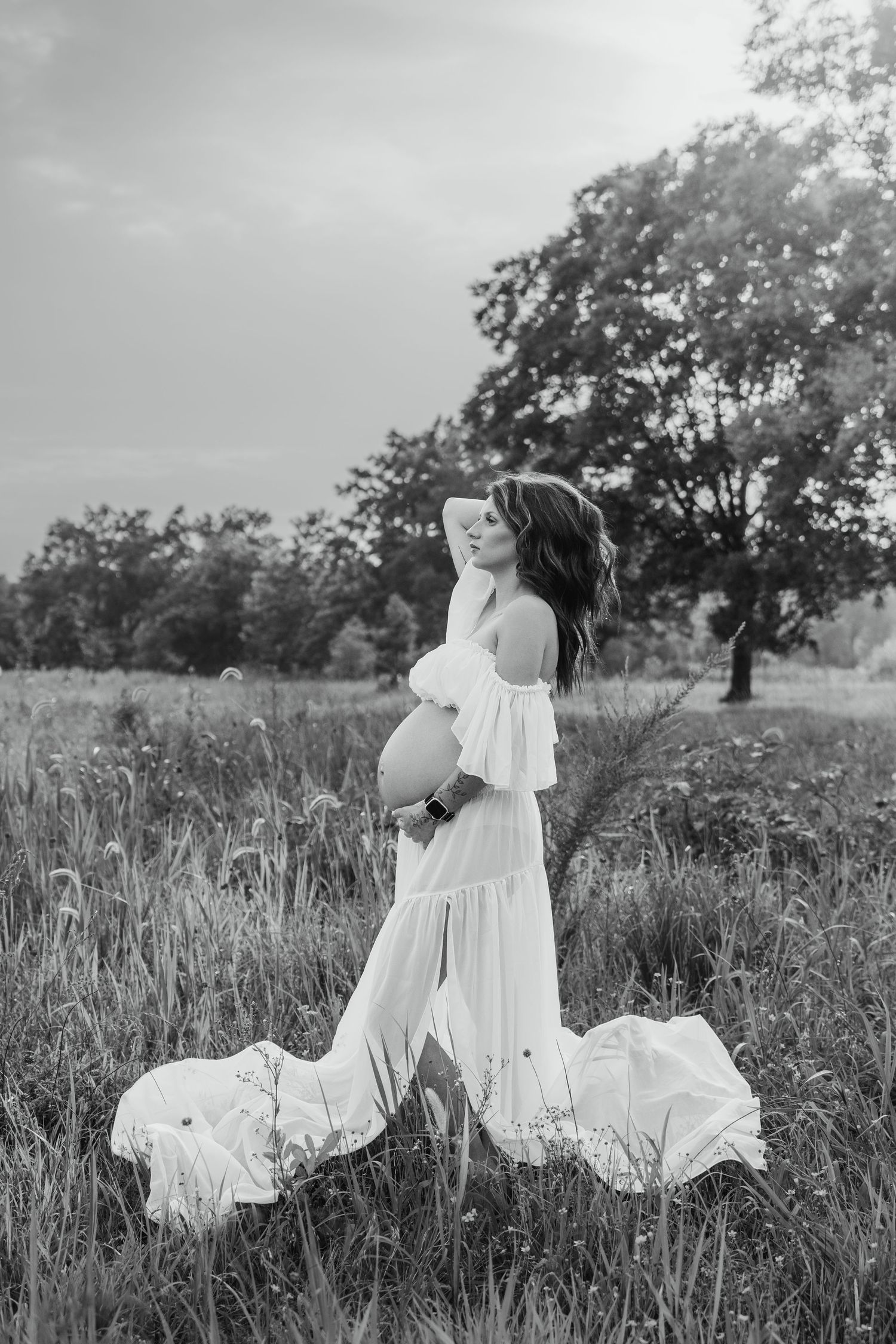 Black and white photo of pregnant woman in white off-shoulder gown standing in field with trees in background.