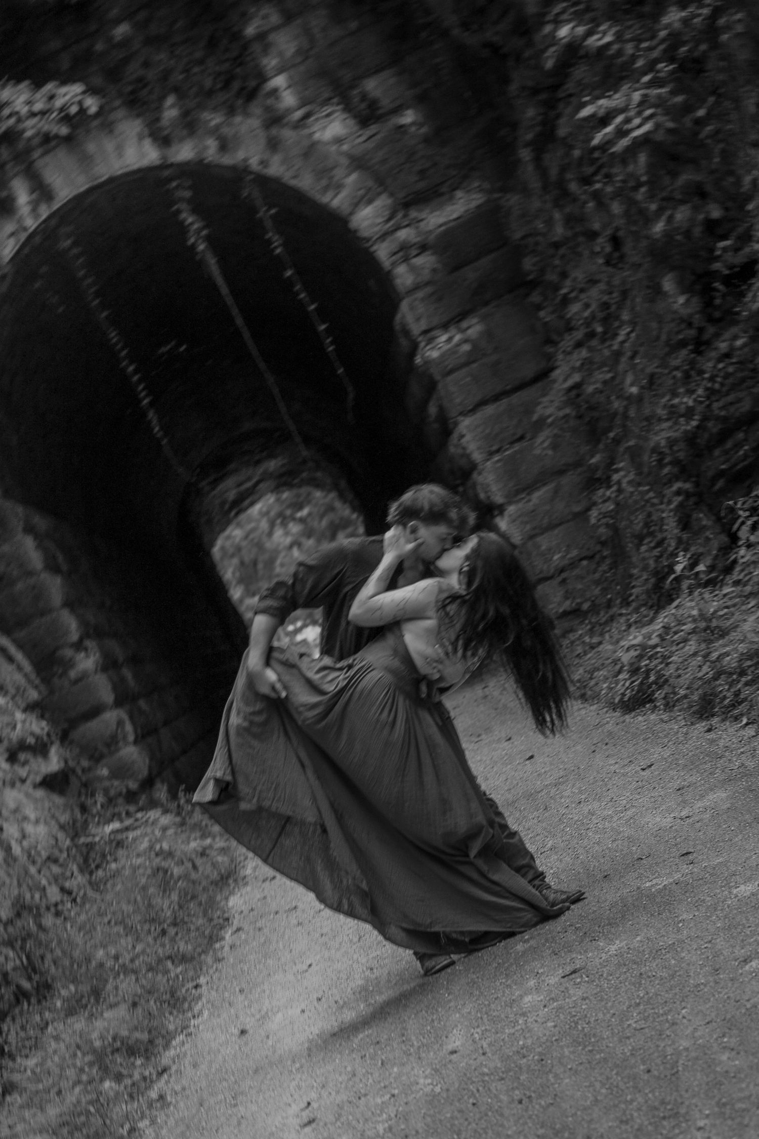 Black and white photo of a couple sharing a passionate kiss and dip in front of a rustic stone tunnel arch.