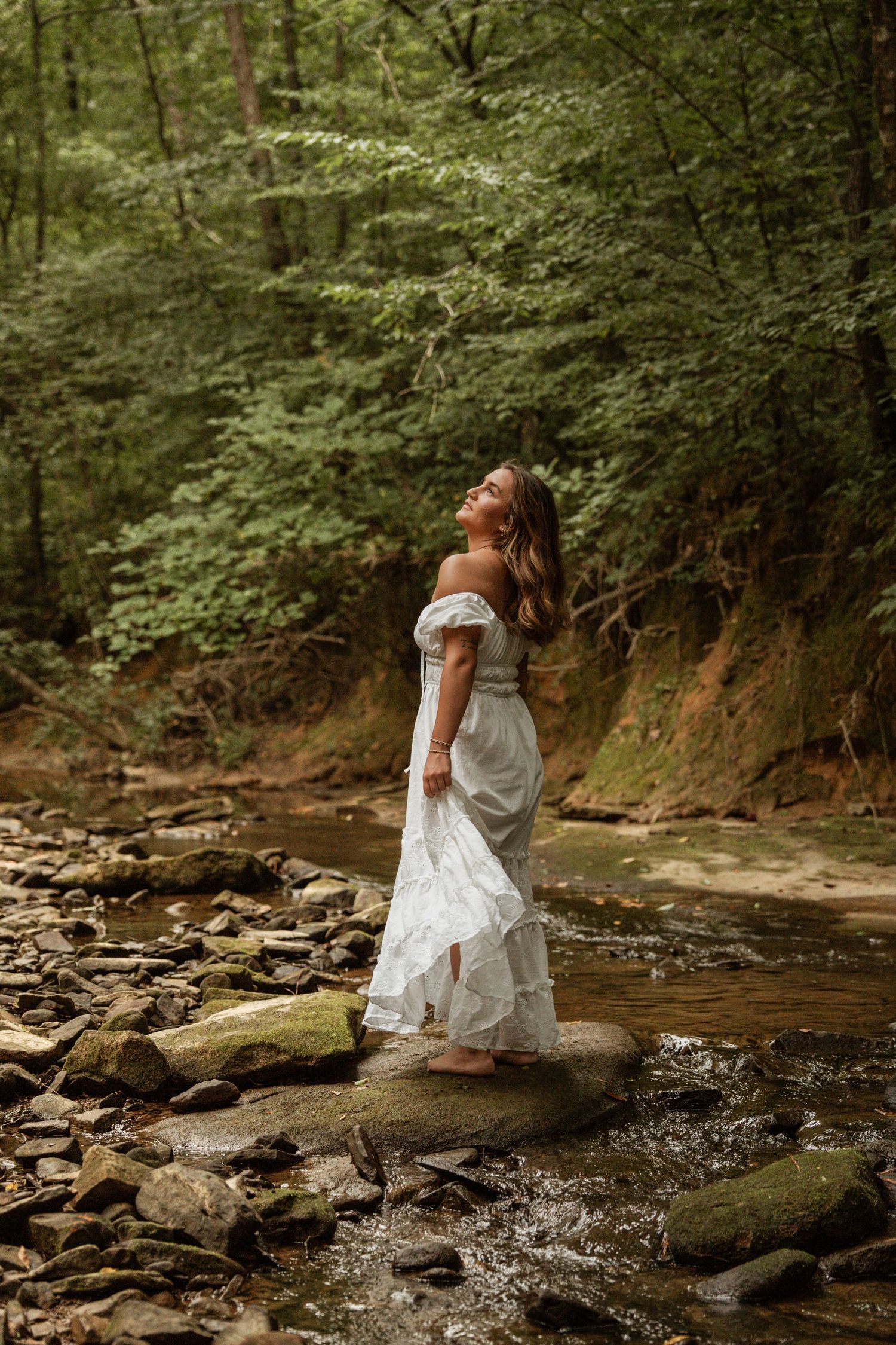 Woman in white off-shoulder dress standing on rocks by a forest creek, gazing upward surrounded by lush green trees.