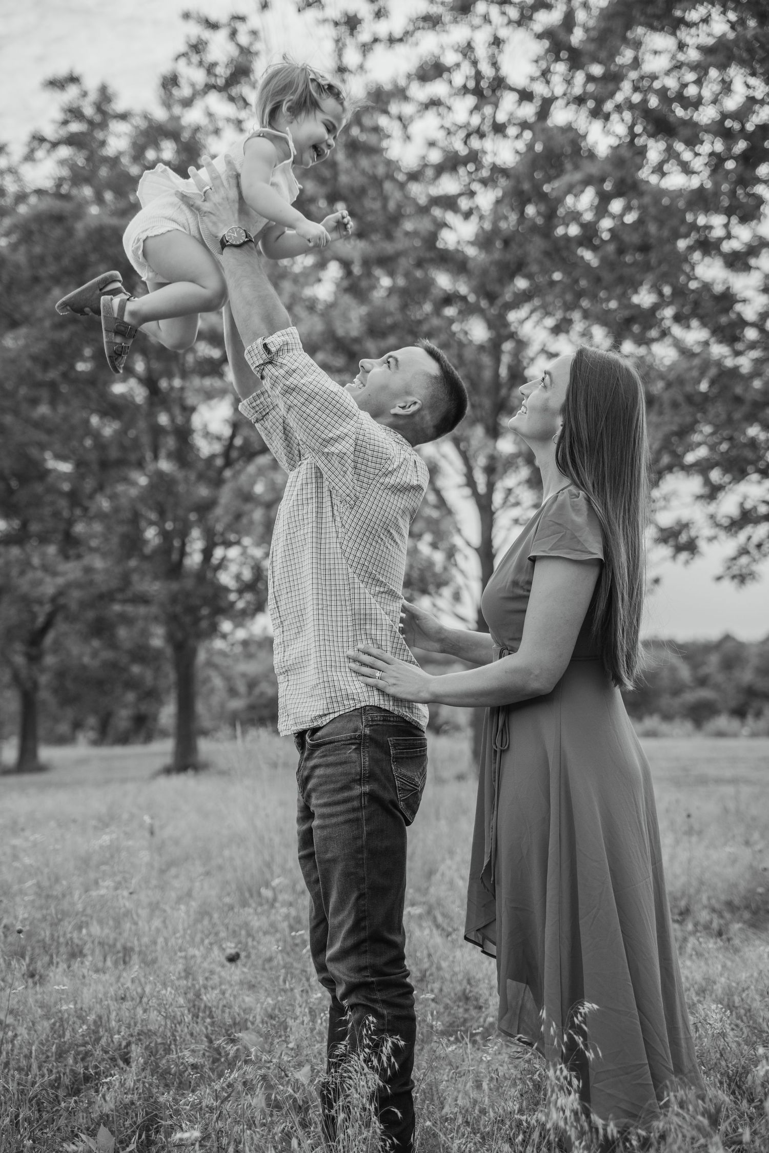 Black and white photo of a father lifting his toddler in the air while mother watches in a park.