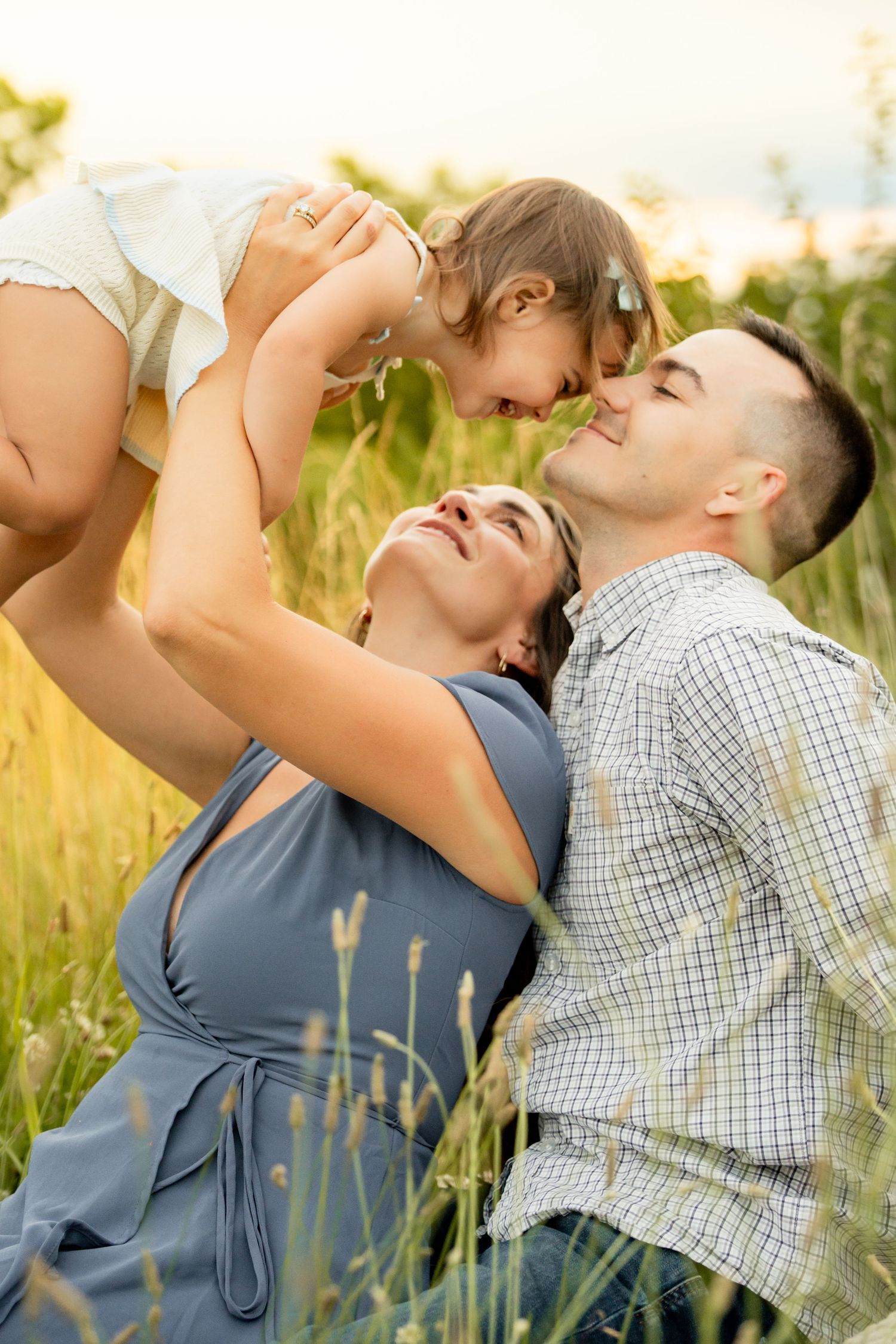 Family of three sitting in tall grass as mother lifts baby girl toward fathers face for a kiss.