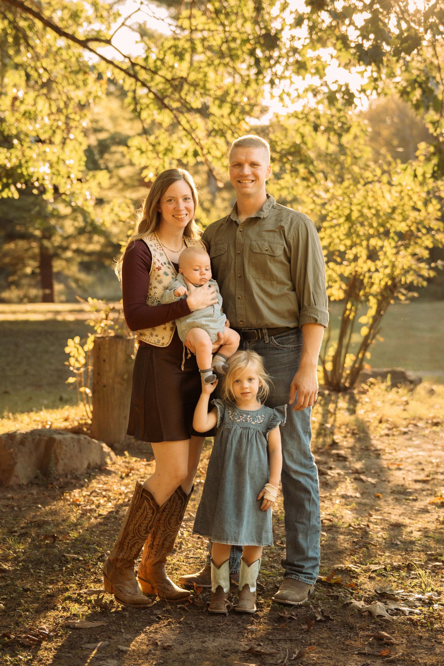 Family of four posing outdoors in golden sunlight, parents holding baby and toddler girl in cowboy boots among lush green trees.