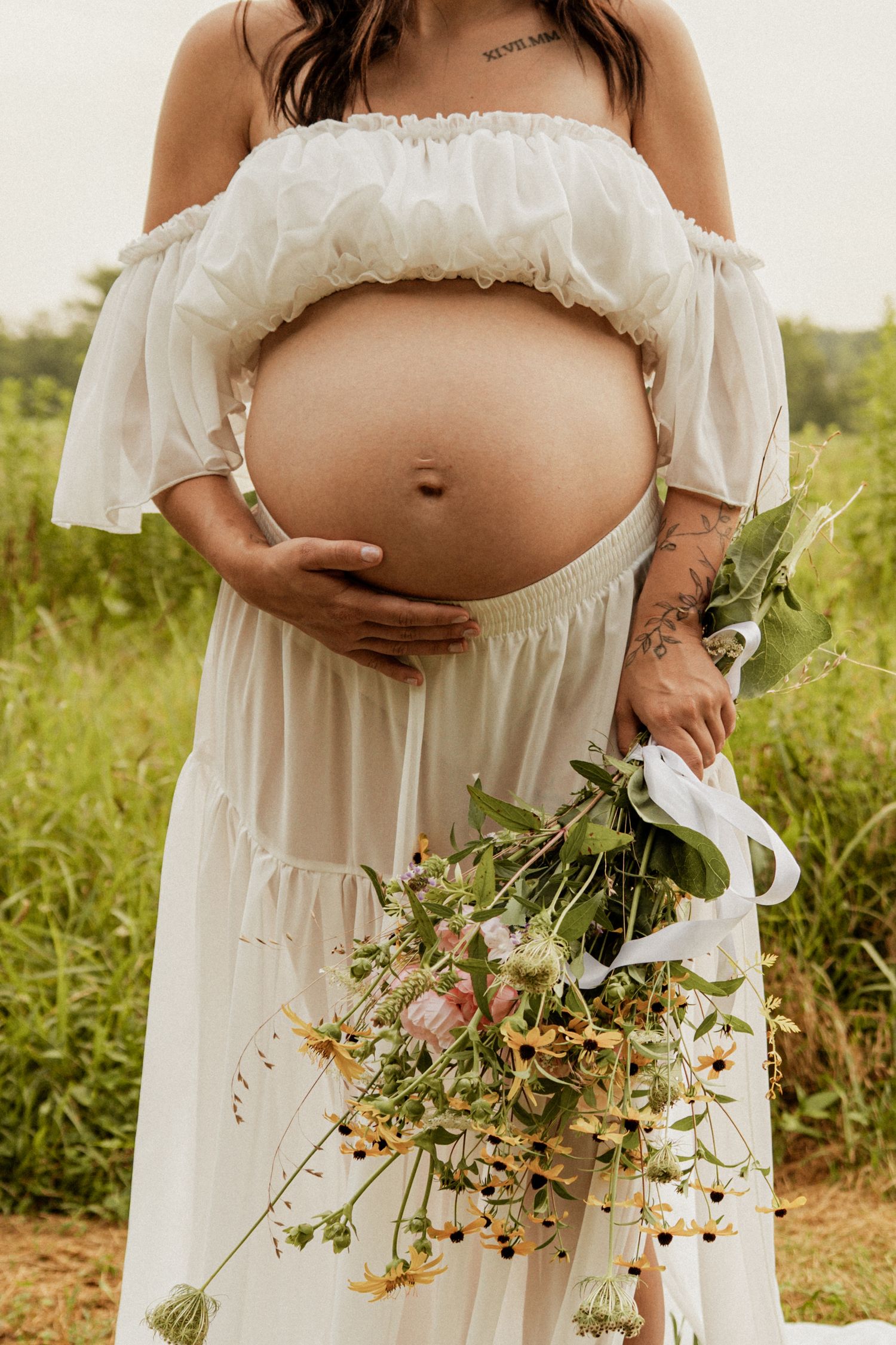 Pregnant woman in a white off-shoulder dress holding a wildflower bouquet while cradling her bump.