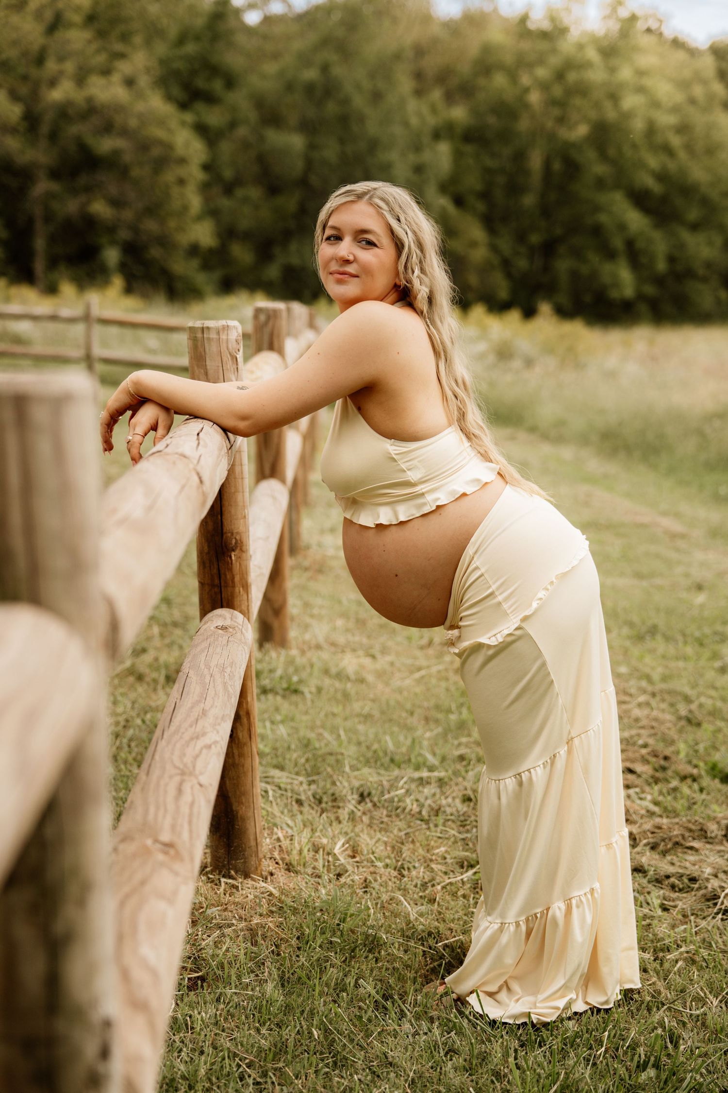 Pregnant woman in cream two-piece outfit leaning on rustic wooden fence in lush green countryside setting.