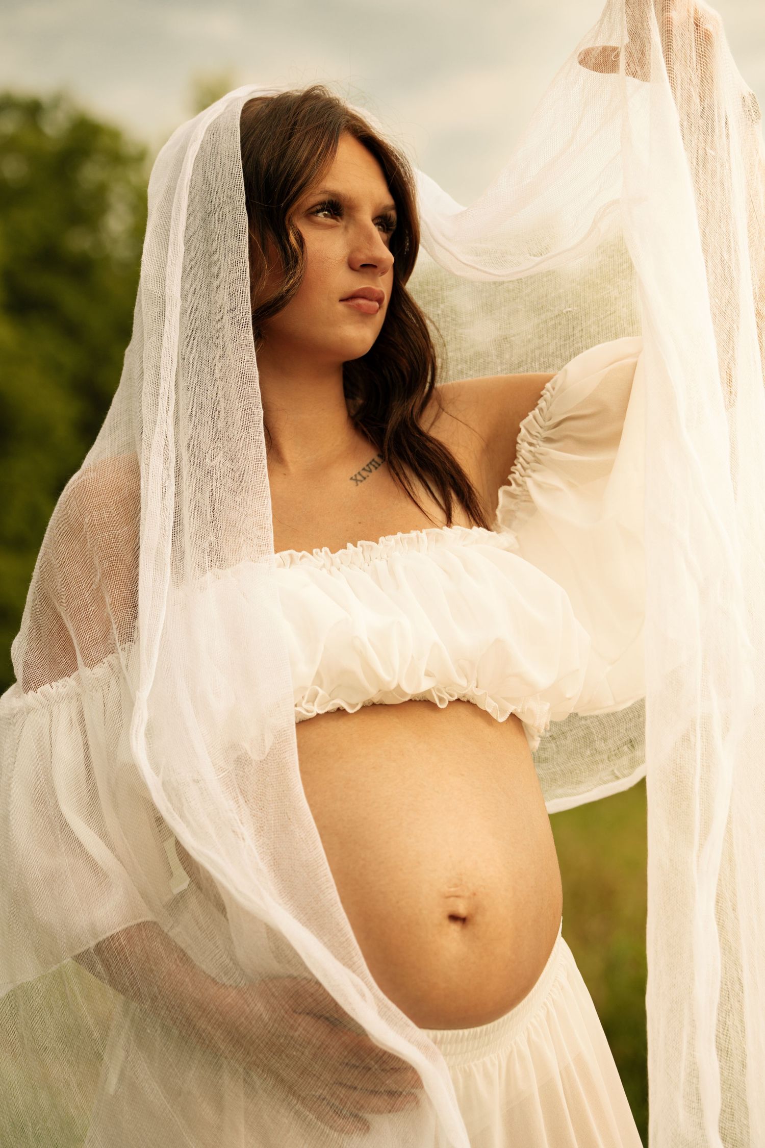 Pregnant woman in white off-shoulder top holds flowing sheer fabric outdoors.