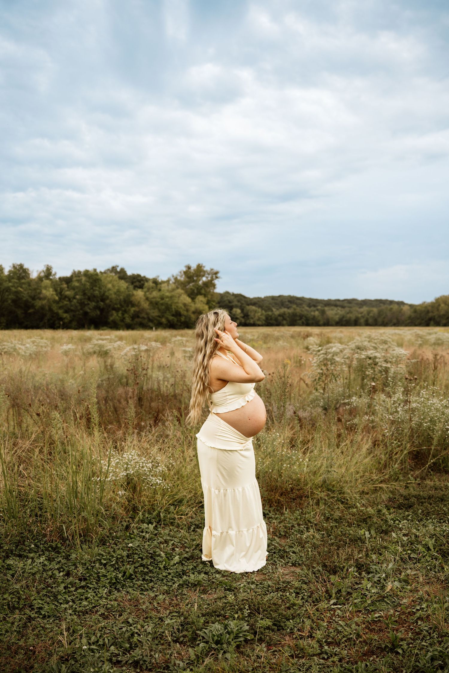 Pregnant woman in white dress standing in a field, cradling her baby bump while looking up at a cloudy sky.
