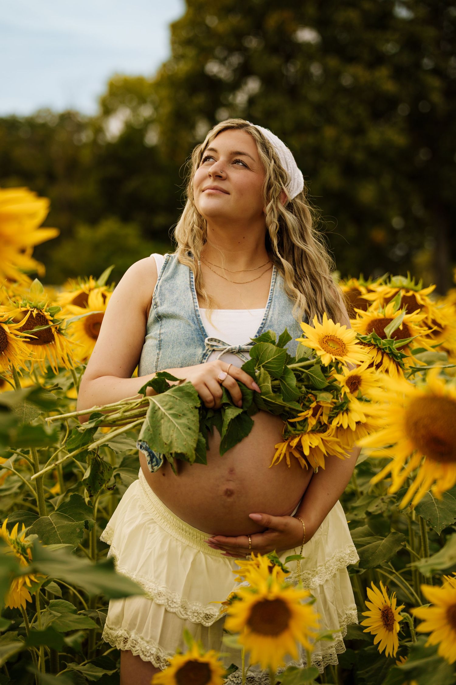 Pregnant woman holding sunflowers in a sunflower field, wearing a denim vest and cream skirt outdoors.