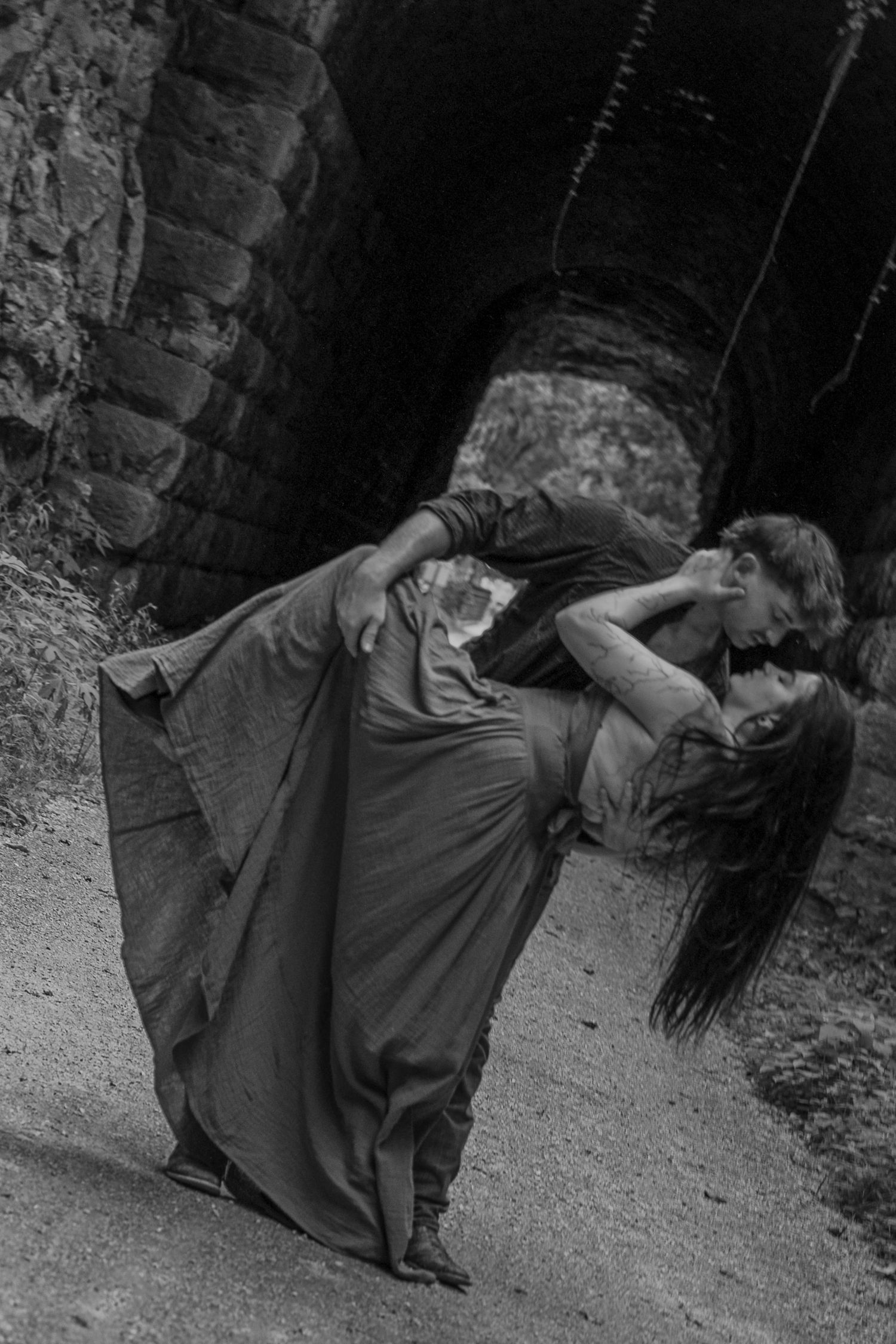 Black and white photo of a couple sharing a passionate dip kiss near a stone tunnel archway.