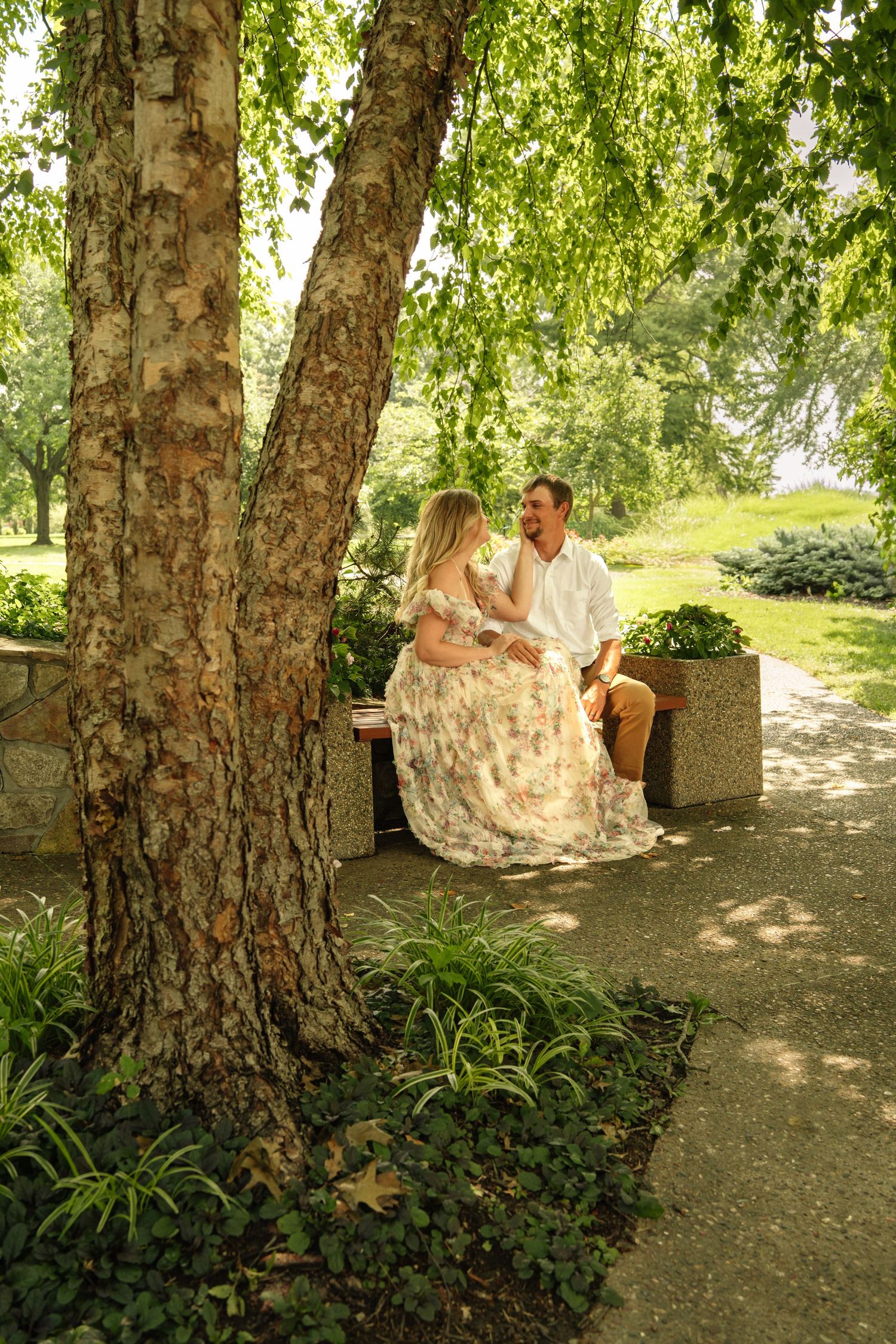 Couple sitting on a bench under a birch tree in a lush green park, woman in floral dress facing man.