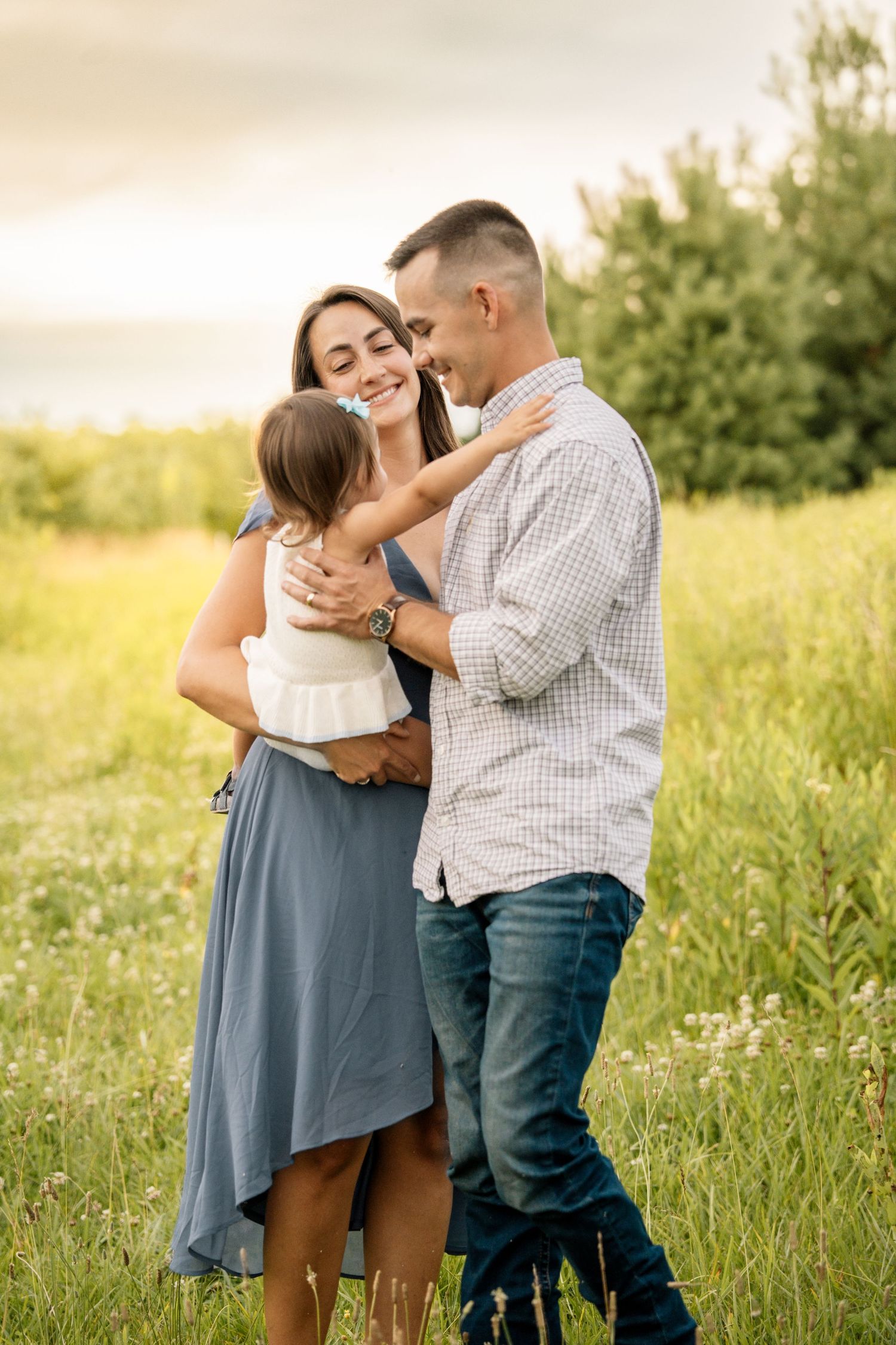 A loving couple shares a tender moment while holding their toddler in a lush green summer field.