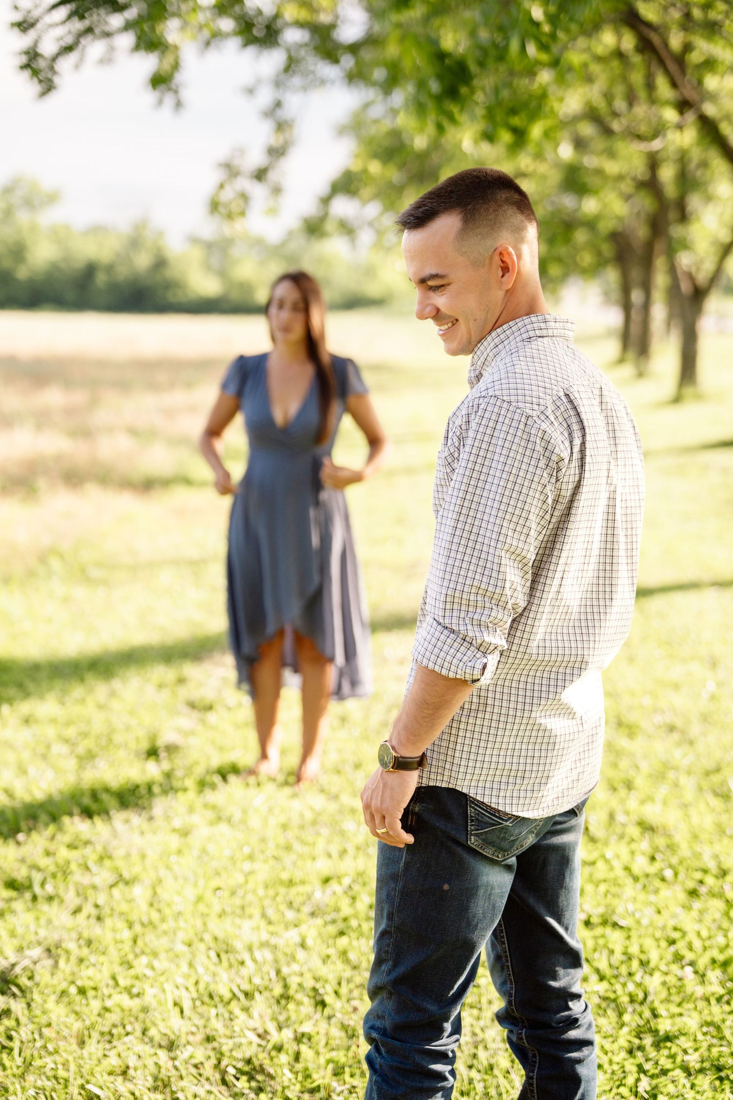 A smiling man in a plaid shirt and jeans stands in a sunny park while a woman in a blue dress stands in the background.