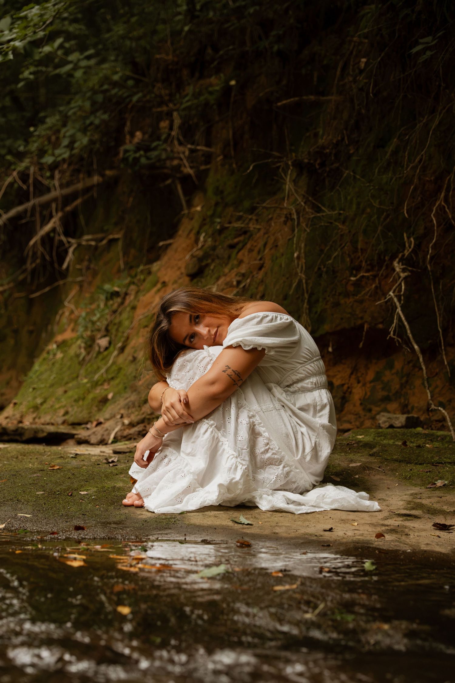 Woman in white dress sits curled up by forest stream, hugging knees near mossy rocks.