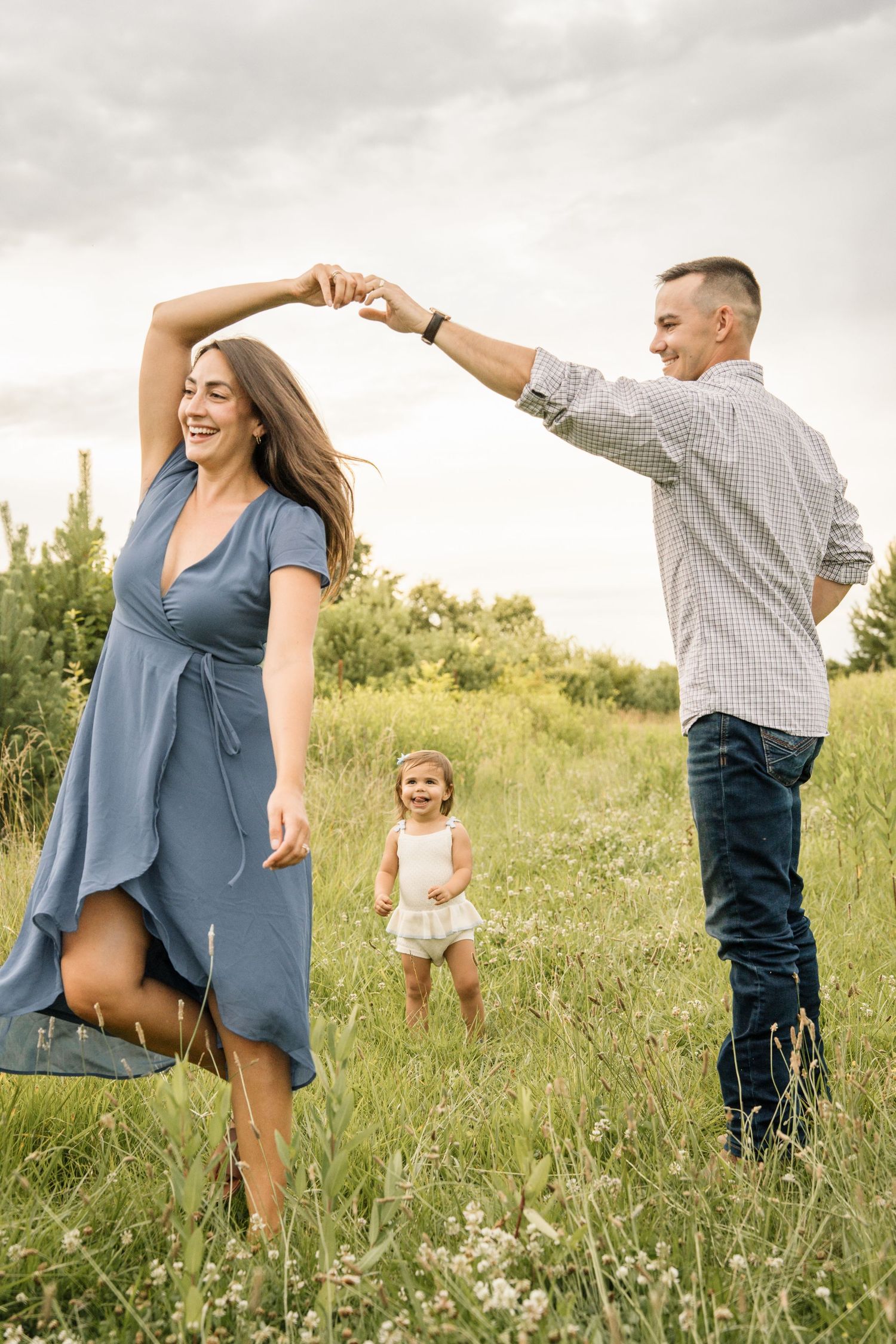 Happy family dancing in green meadow, father spinning mother while toddler watches joyfully.