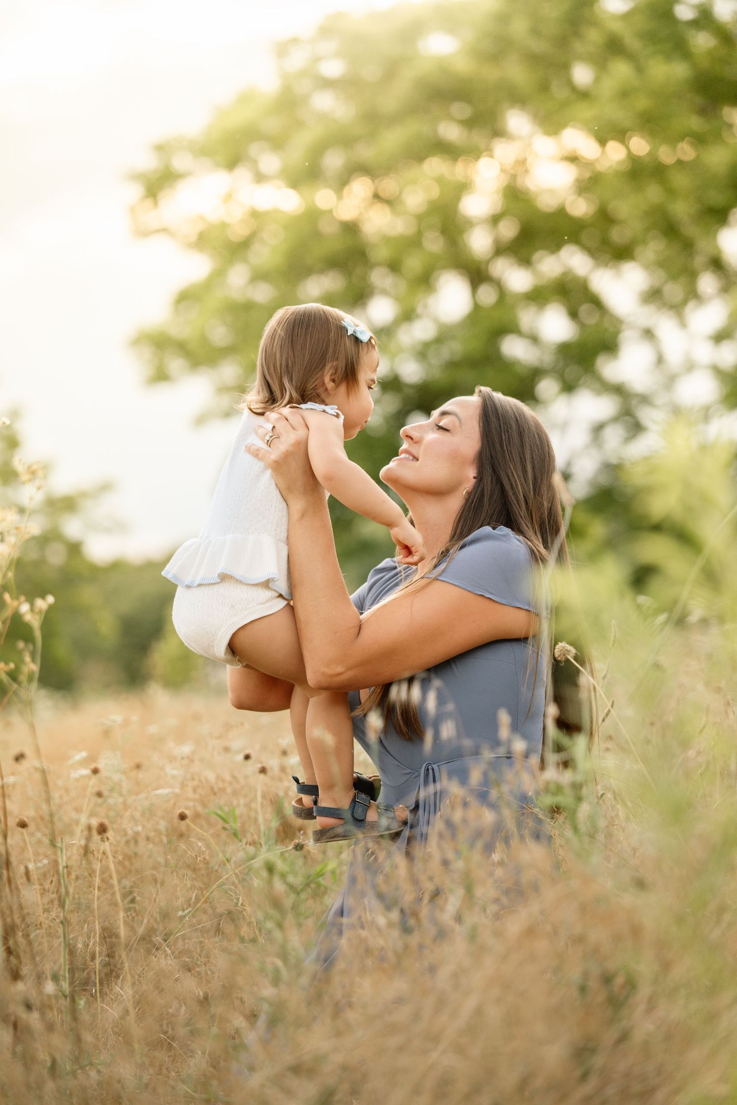 Mother sitting in golden grass field lifting her toddler daughter up close for a nose kiss.