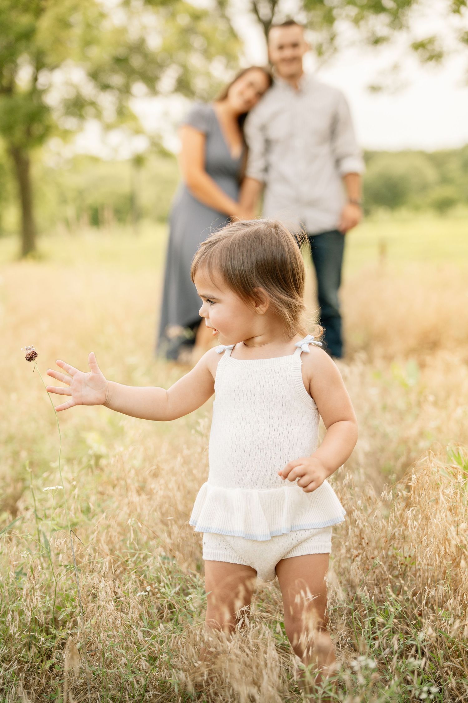 Toddler girl in white romper plays in golden field while parents embrace in soft focus background.