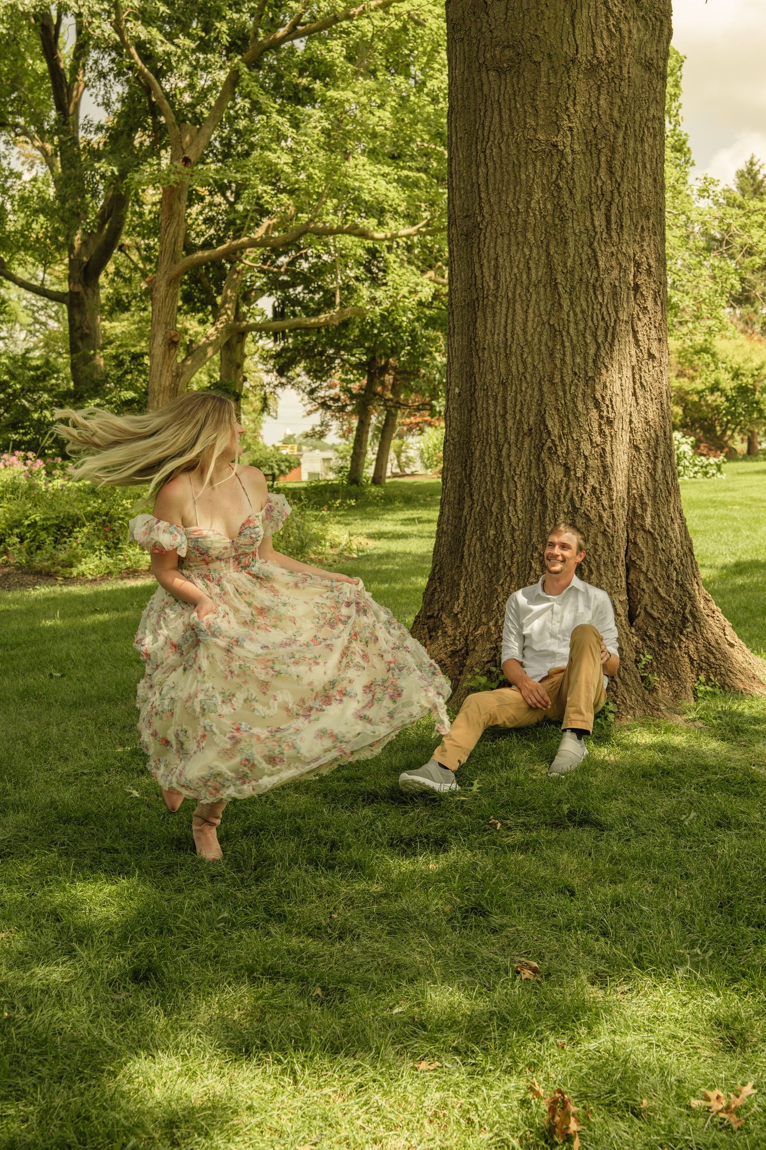 Woman in floral dress twirling near large tree while man sits on grass smiling in sunny green park.