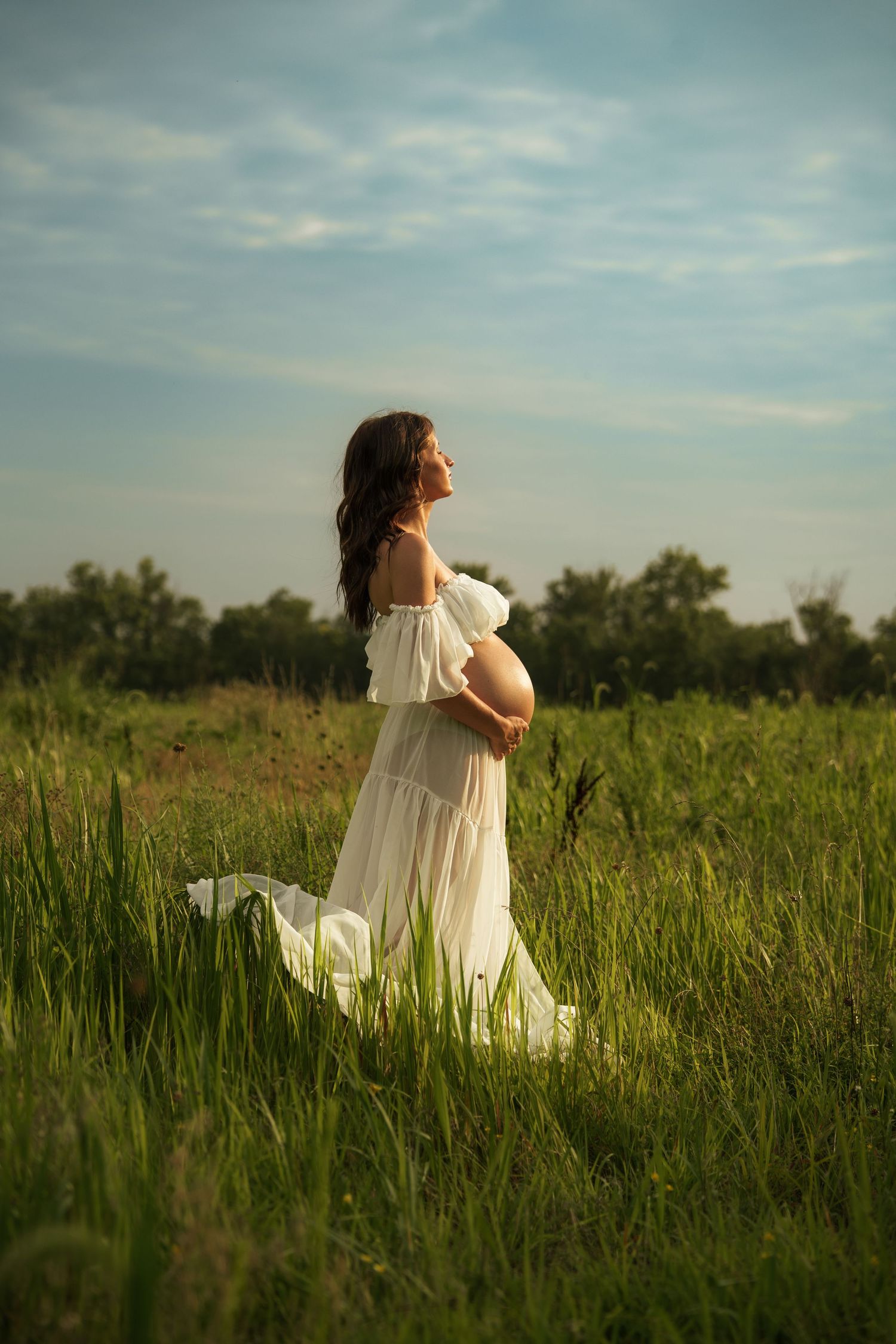 Pregnant woman in white off-shoulder dress standing in green field, cradling baby bump, with blue sky backdrop.