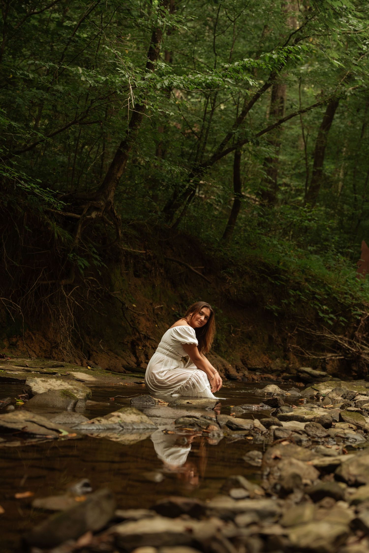 Woman in white dress sitting in a rocky forest creek with reflection in the water.