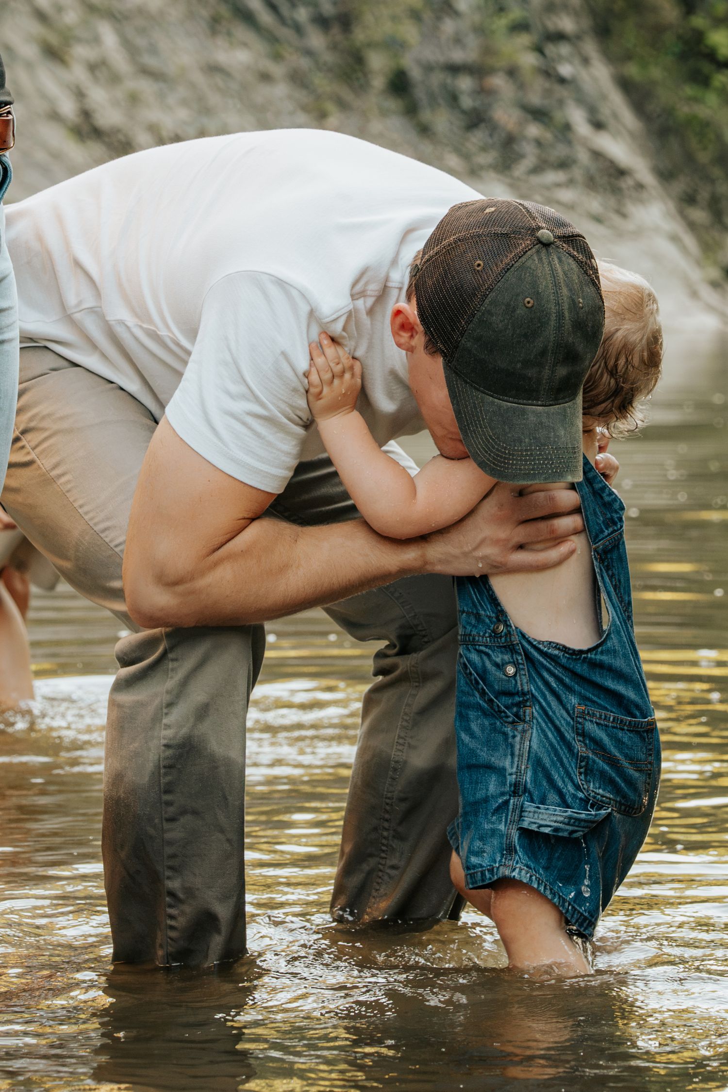 A father bends down to embrace his toddler in denim overalls while wading in a shallow river.