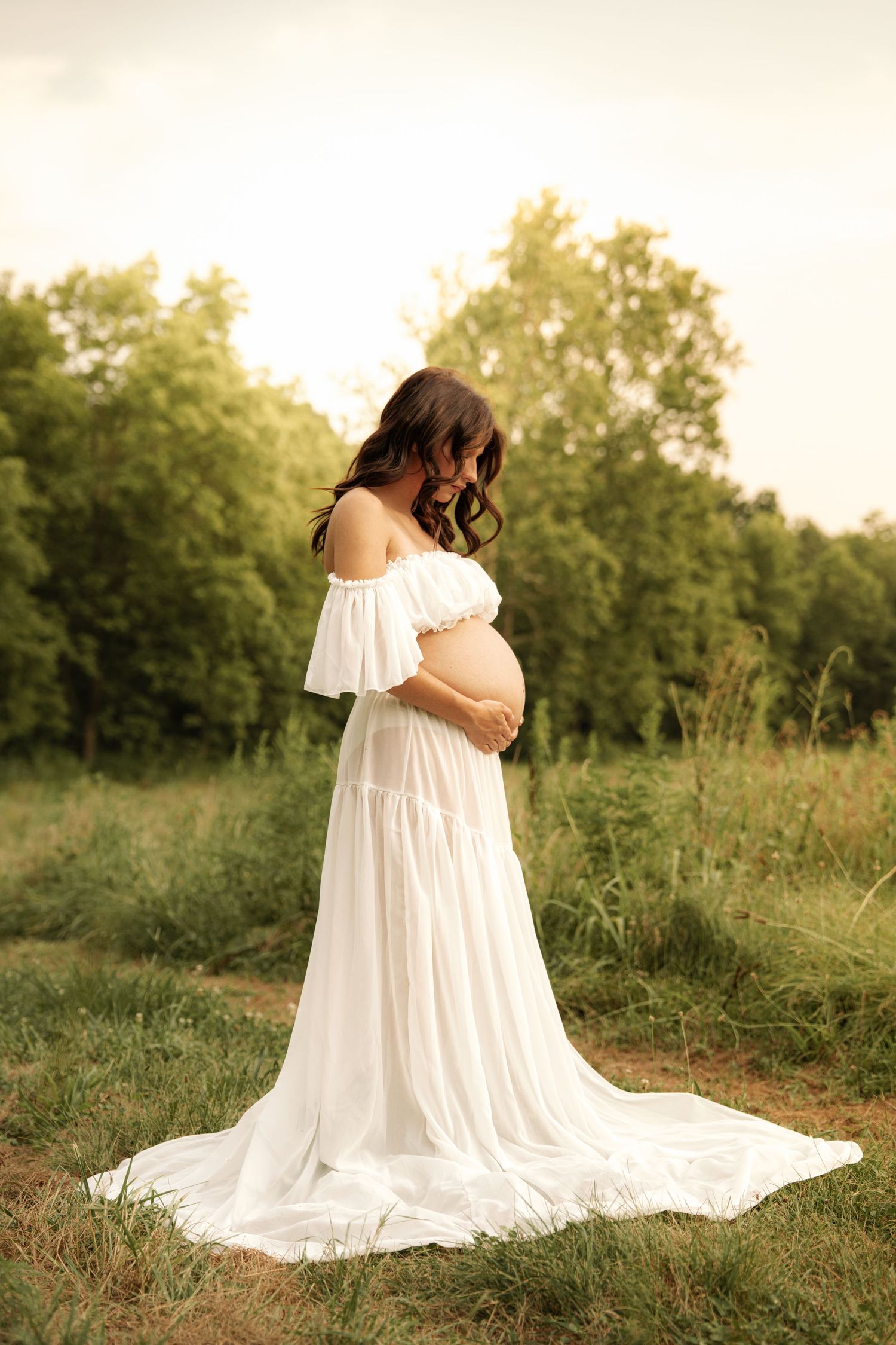 Pregnant woman in a white off-shoulder gown cradling her baby bump in a scenic outdoor field.