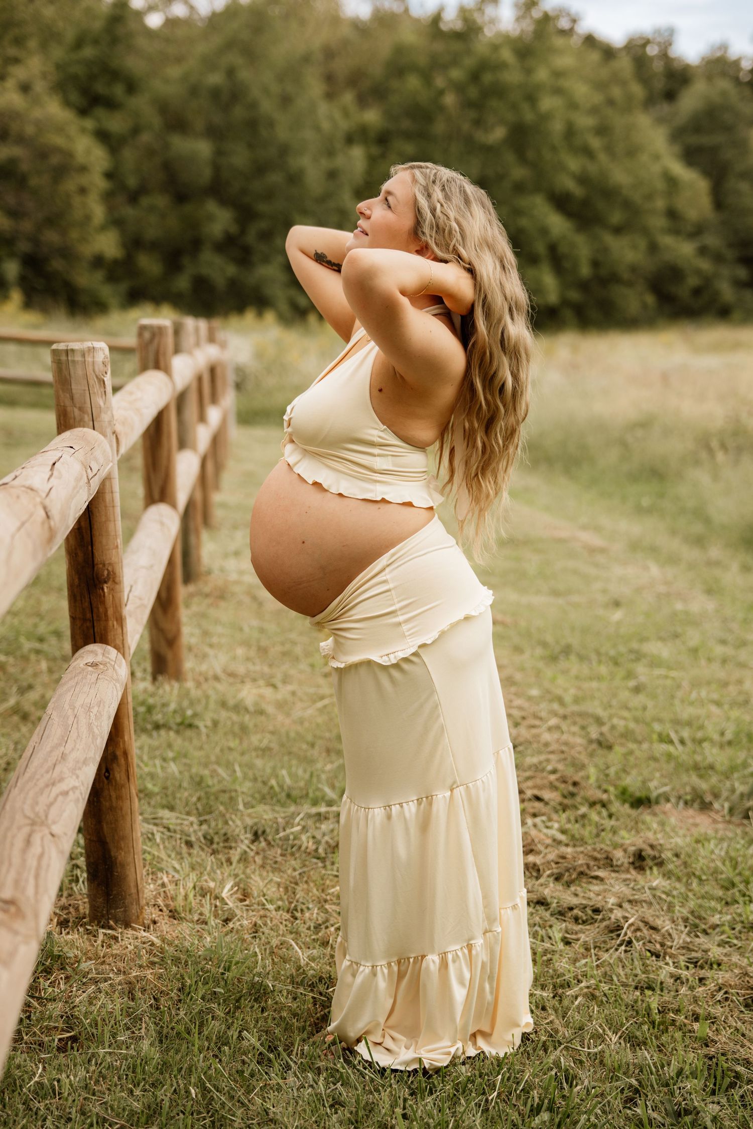 Pregnant woman in cream crop top and maxi skirt posing by rustic wooden fence in green field.