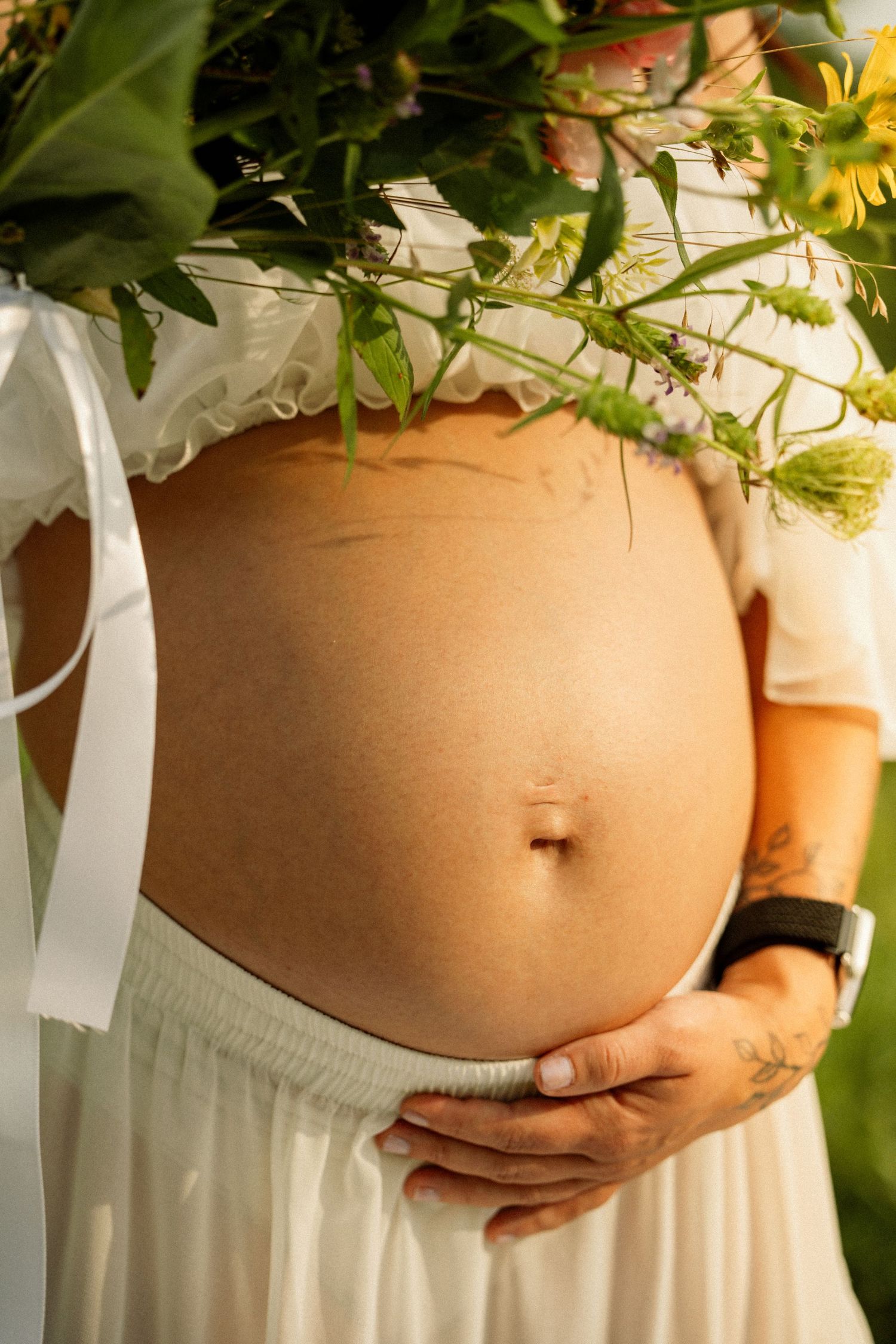Close-up of a pregnant belly with a white dress and wildflower bouquet overhead.