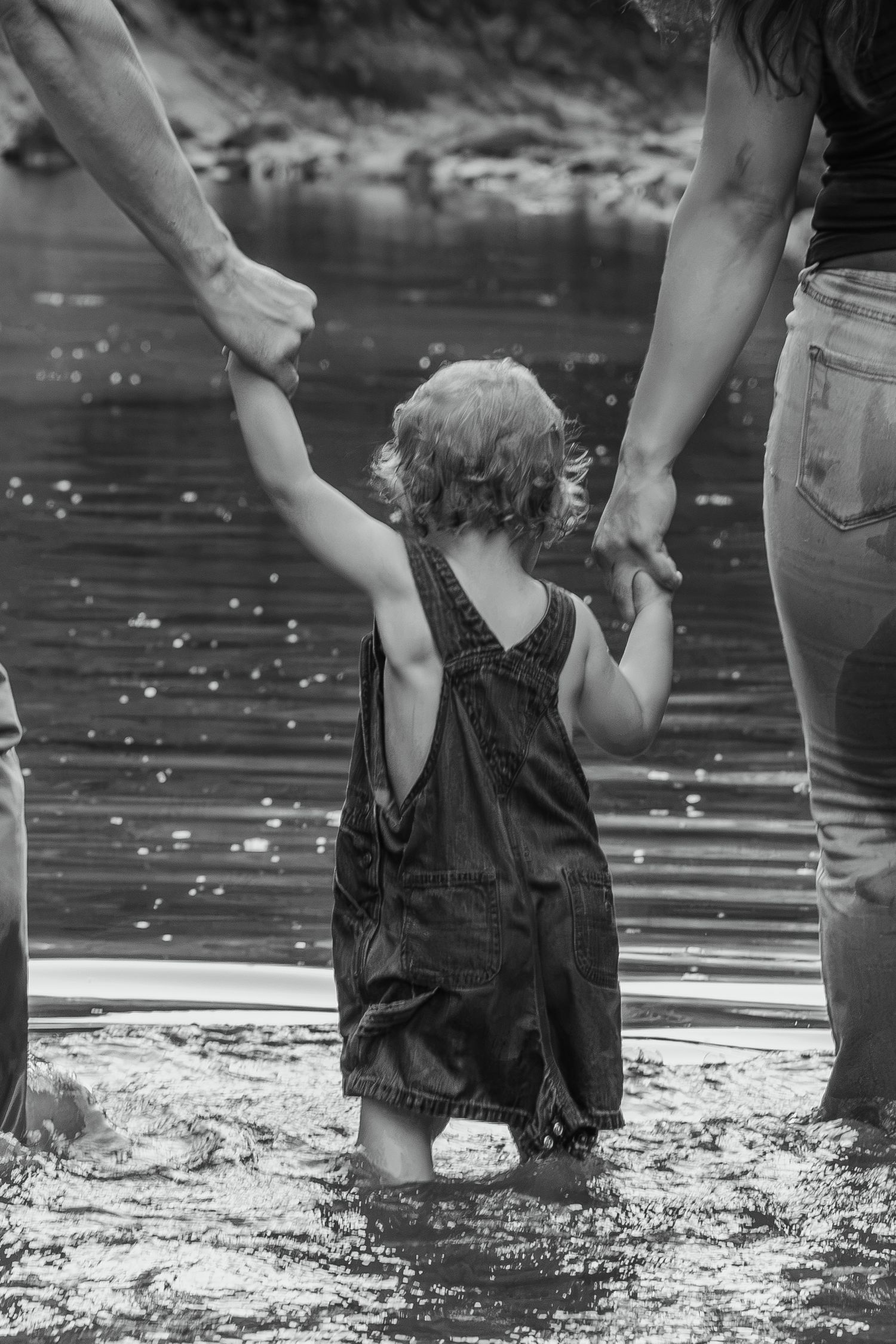 Black and white photo of a toddler in overalls wading in water, holding hands with two adults on either side.