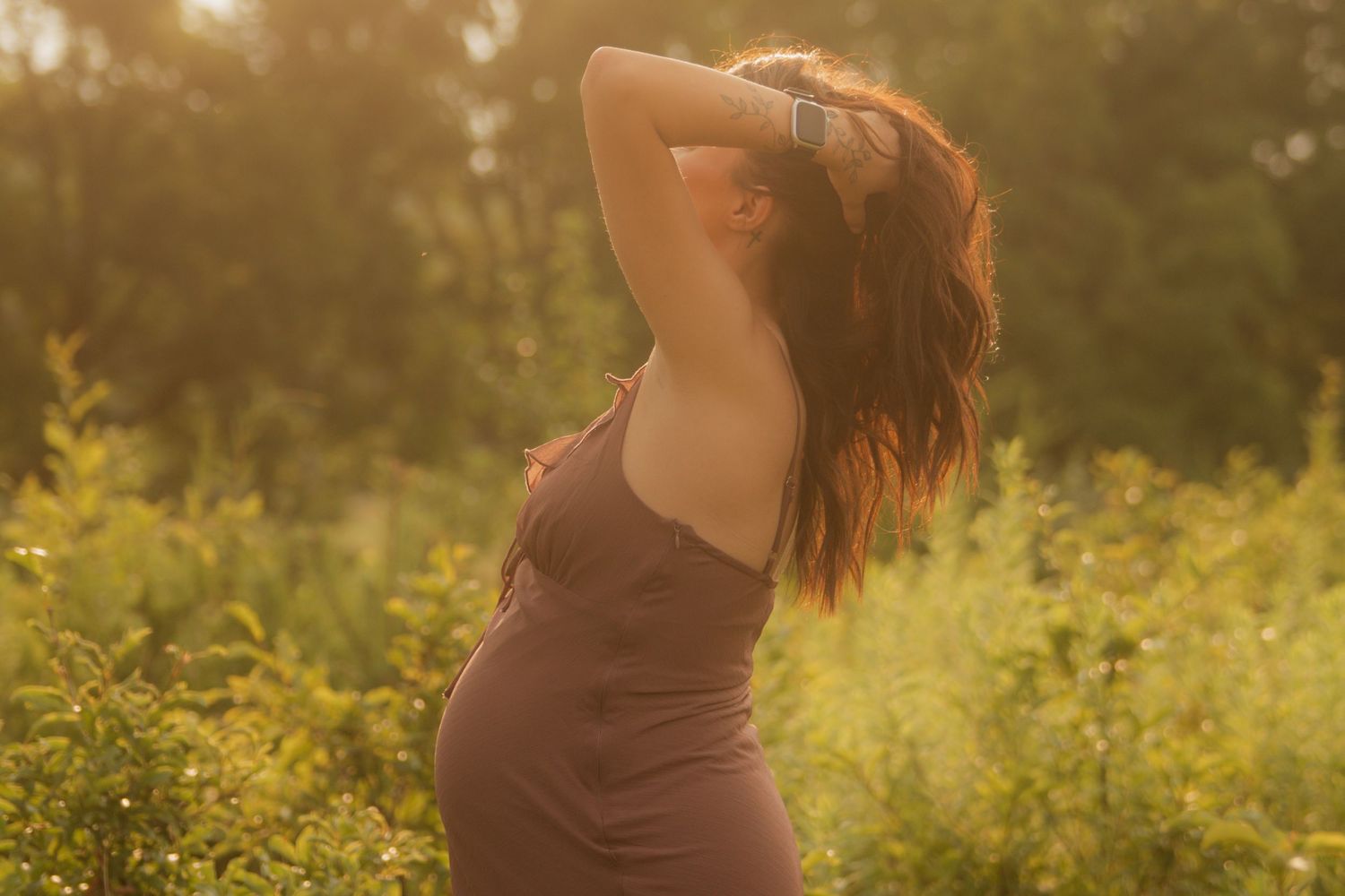 Pregnant woman in brown dress poses outdoors in golden sunlight surrounded by lush green foliage.