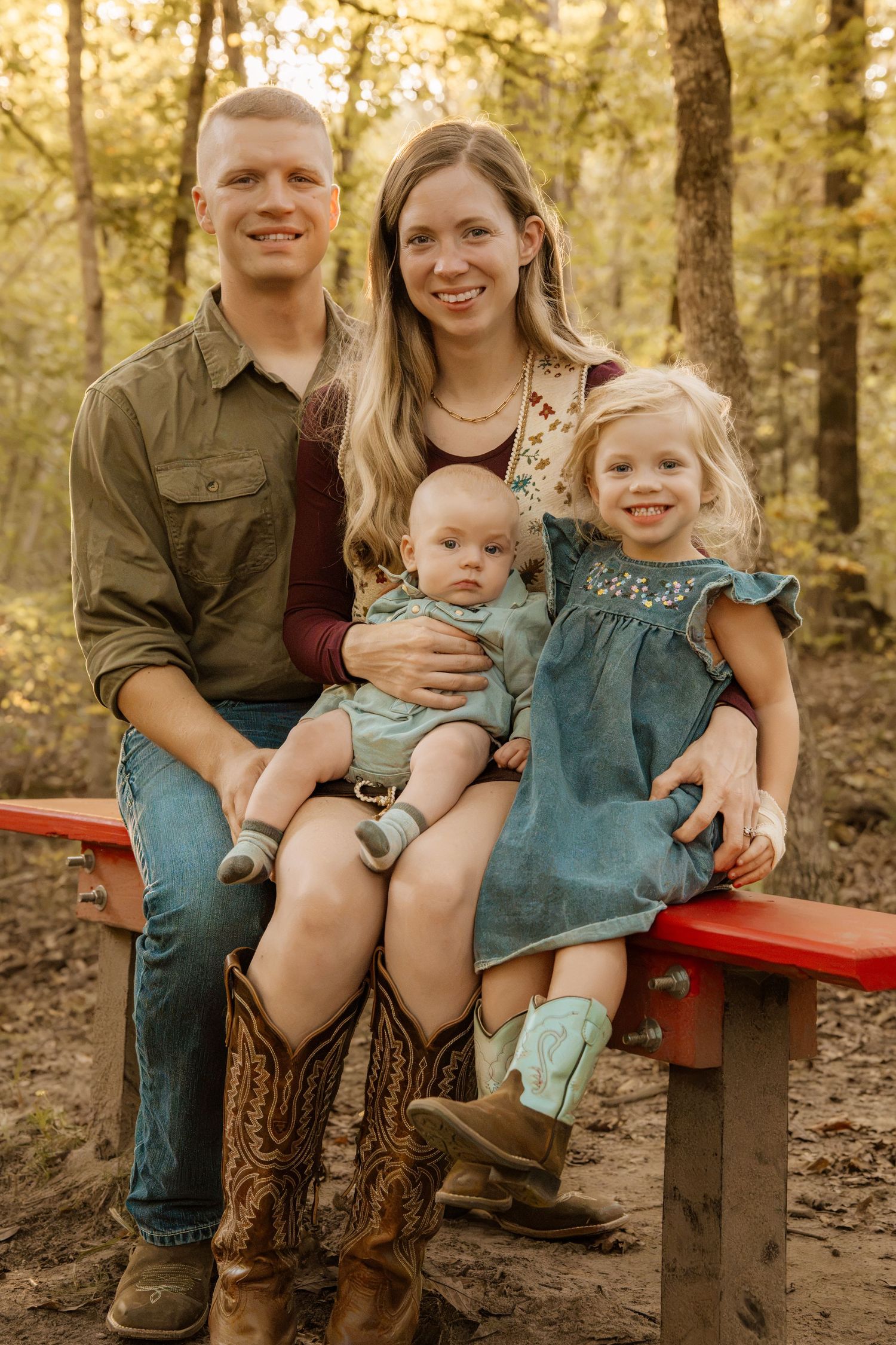 Happy family of four posing on a red bench in an autumn forest, parents with baby and toddler girl.