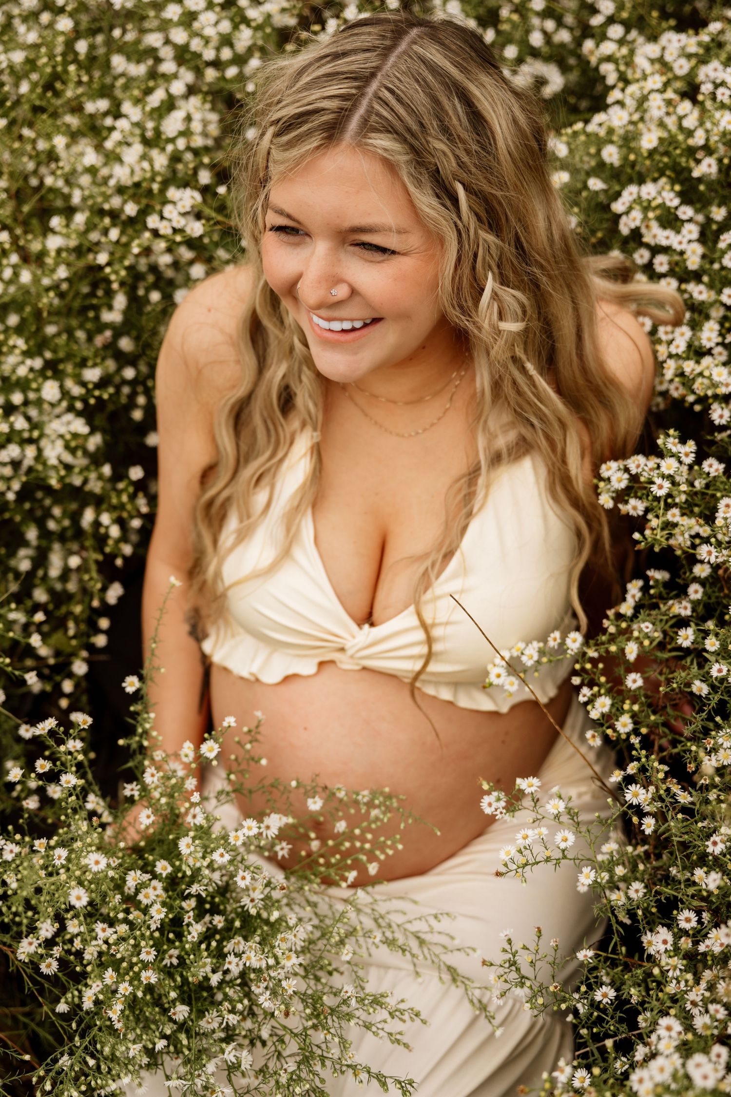 Joyful pregnant woman in cream ruffled top sits surrounded by delicate white wildflowers laughing.