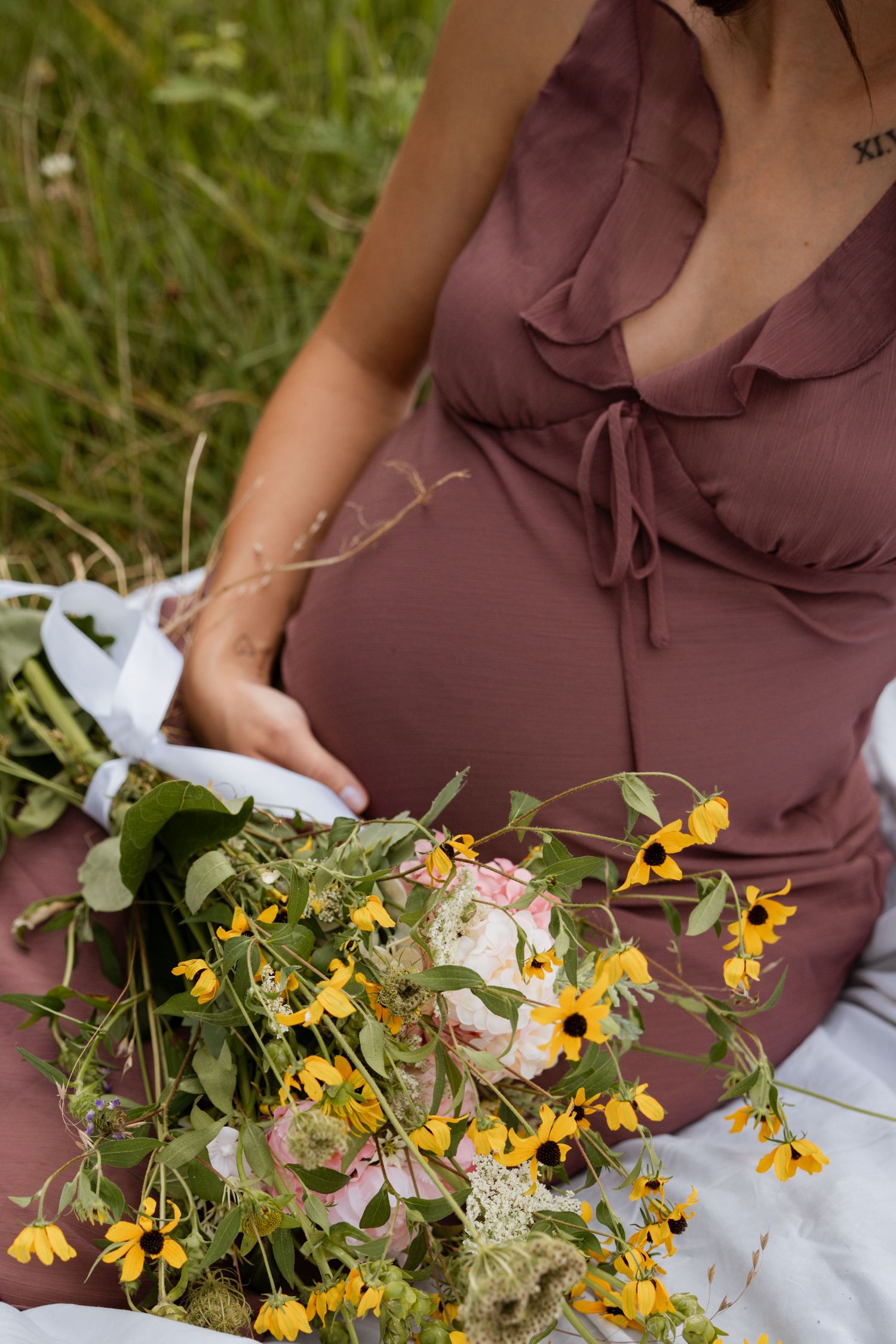 Pregnant woman in mauve dress sitting in grass holding wildflower bouquet with yellow and white blooms.
